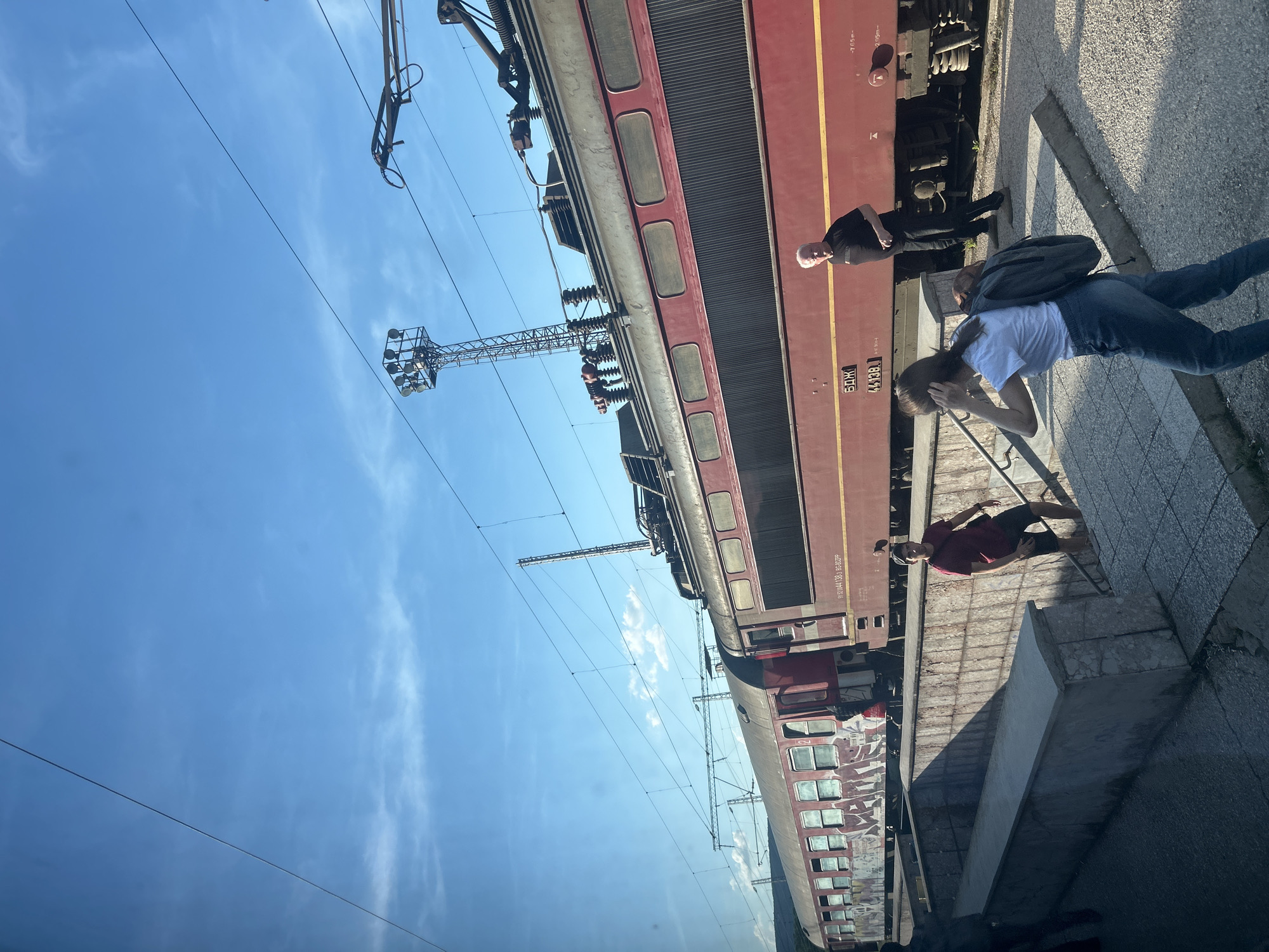 The image captures a scene at a train station in Kochovo, Bulgaria, under a clear blue sky with scattered clouds. A long, red train with graffiti on its side is stationary on the tracks. In the foreground, a set of concrete stairs leads down from ground level. A young woman with a backpack walks up the stairs, facing away from the camera, while a man in a red shirt and dark shorts also ascends the stairs, looking towards the train. Further back, near the train, an older man with white hair and a black t-shirt stands facing the train. Overhead, power lines and a tall electrical pylon are visible, indicating the presence of an electrified railway. The overall environment suggests a functional, outdoor train station setting.