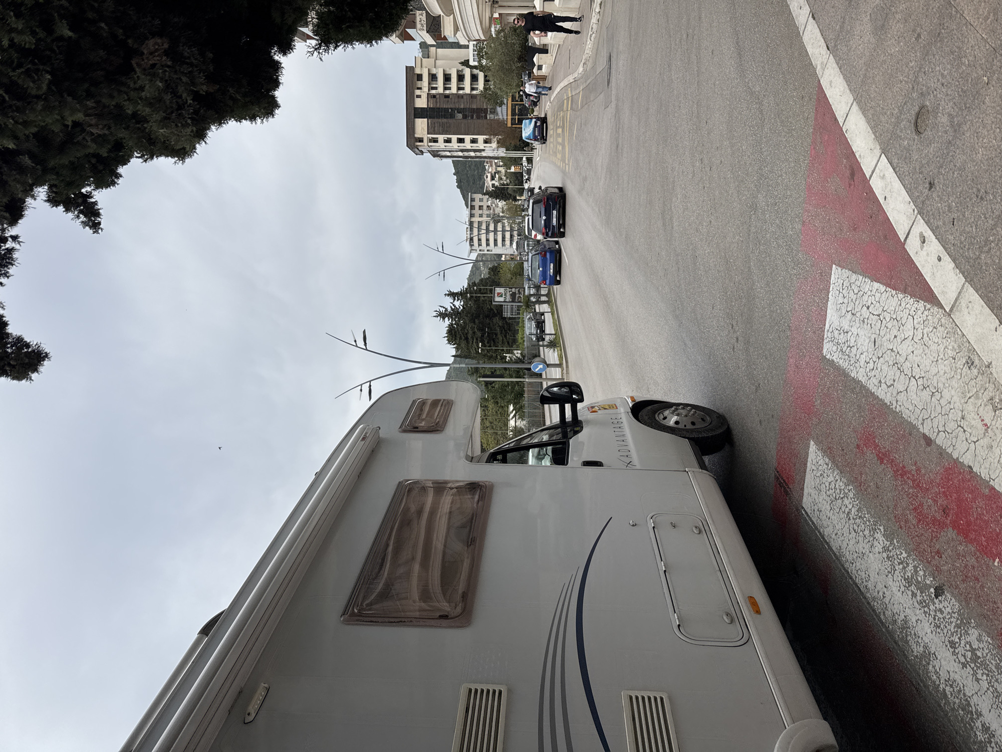 The scene depicts a street in Budva, Montenegro, with a white camper van parked along the sidewalk on a red and white marked zone. The vehicle is positioned on the right side of the image, with its side facing the viewer. Cars are parked along the street, and buildings and trees populate the background. Pedestrians are visible on the sidewalk further down the street. The sky is overcast.