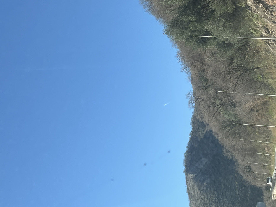 A panoramic landscape photograph, reportedly captured in Brežine, Montenegro, depicts a mountainous road scene under a clear sky. The upper two-thirds of the frame are dominated by a cloudless, light blue sky. Several dark, irregular smudges are visible in the upper left quadrant, consistent with dust or imperfections on a camera lens or viewing surface. A faint, white contrail, indicative of an aircraft's passage, is present in the upper right, arcing subtly downwards.
The lower third of the image features rugged hills and mountains, their slopes covered with sparse vegetation. Numerous trees appear deciduous and barren, while some sections exhibit green evergreen foliage, particularly on the right side. Along the base of the mountains, a paved road is visible in the lower left portion of the image. A white vehicle, possibly a van or car, is partially visible on this road, moving away from the viewer. Multiple slender, light-colored poles, consistent with utility or street light poles, are evenly spaced along the roadside, extending vertically into the midground from the lower center and right. No individuals or direct interactions are discernible within the scene.