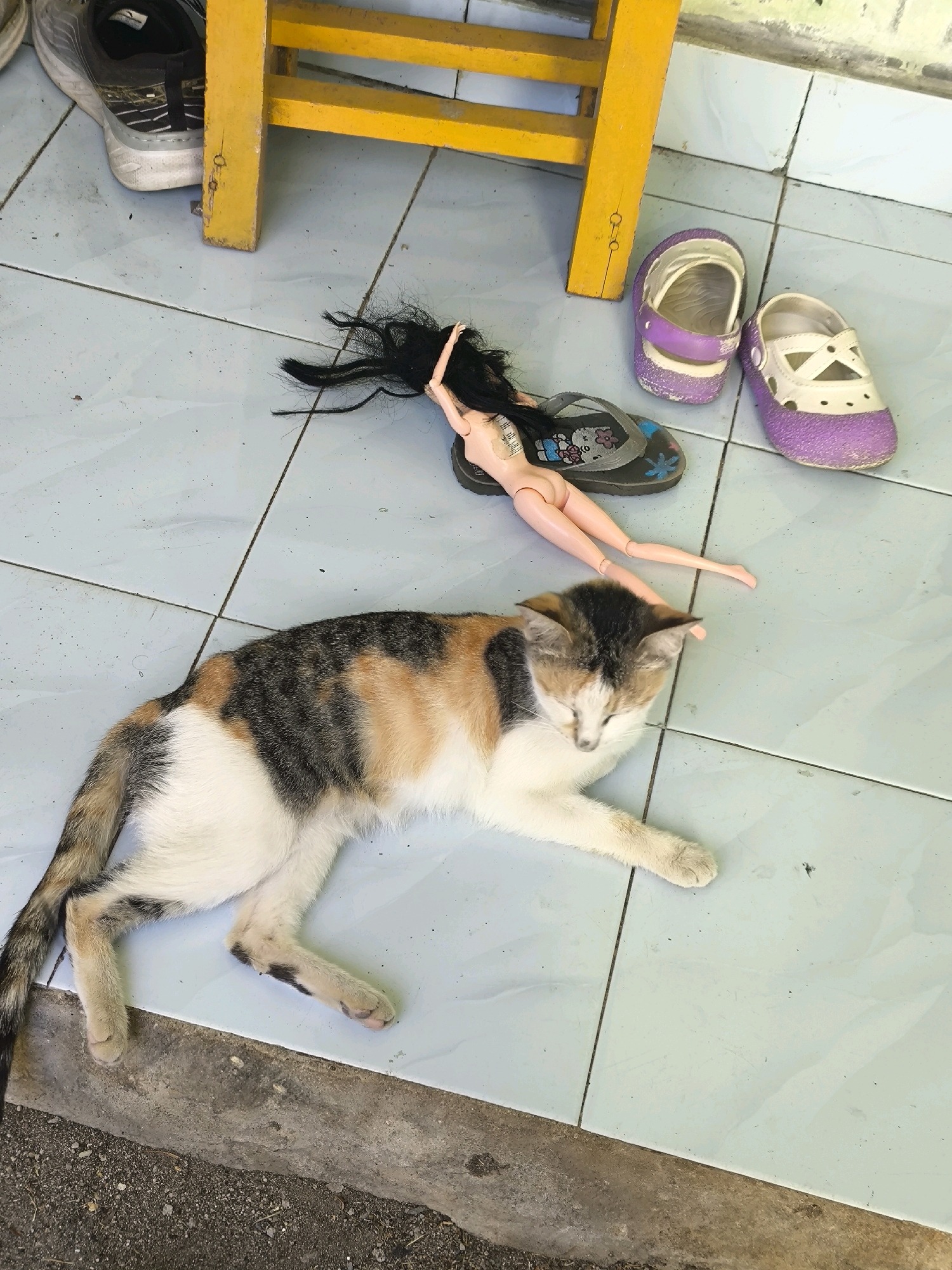A calico cat is sleeping on a light blue tiled floor next to a yellow stool. To the right of the cat, a doll is lying on a gray flip-flop. Two pairs of children's sandals, one purple and white and the other white and purple, are placed near the doll. A gray and white shoe is visible in the upper left corner, partially under the yellow stool. The scene appears to be outdoors, possibly in an urban environment, given the tiled flooring and a concrete border in the lower part of the image. The lighting suggests it is daytime.