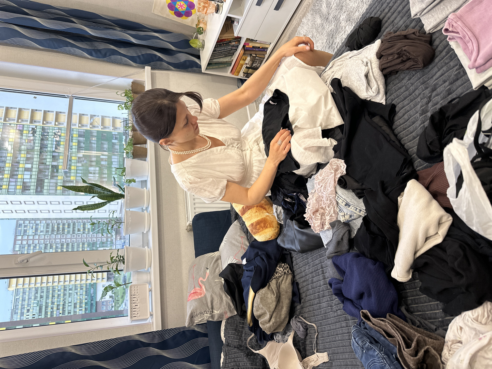 A woman wearing a white blouse and a pearl necklace sits amidst a large pile of clothes on a bed with a gray quilted comforter. The clothes are a mix of colors and fabrics, including white, black, gray, and blue garments. A loaf of bread is also part of the pile. The room is lit by natural light coming from a window to the left, where plants in white pots are displayed. Outside the window, a tall apartment building with a repeating pattern of windows is visible. To the right of the woman, there is a white shelf unit with books and decorative items. Based on the style of the buildings and the decor, the location is likely a modern apartment in Murino, Russia.