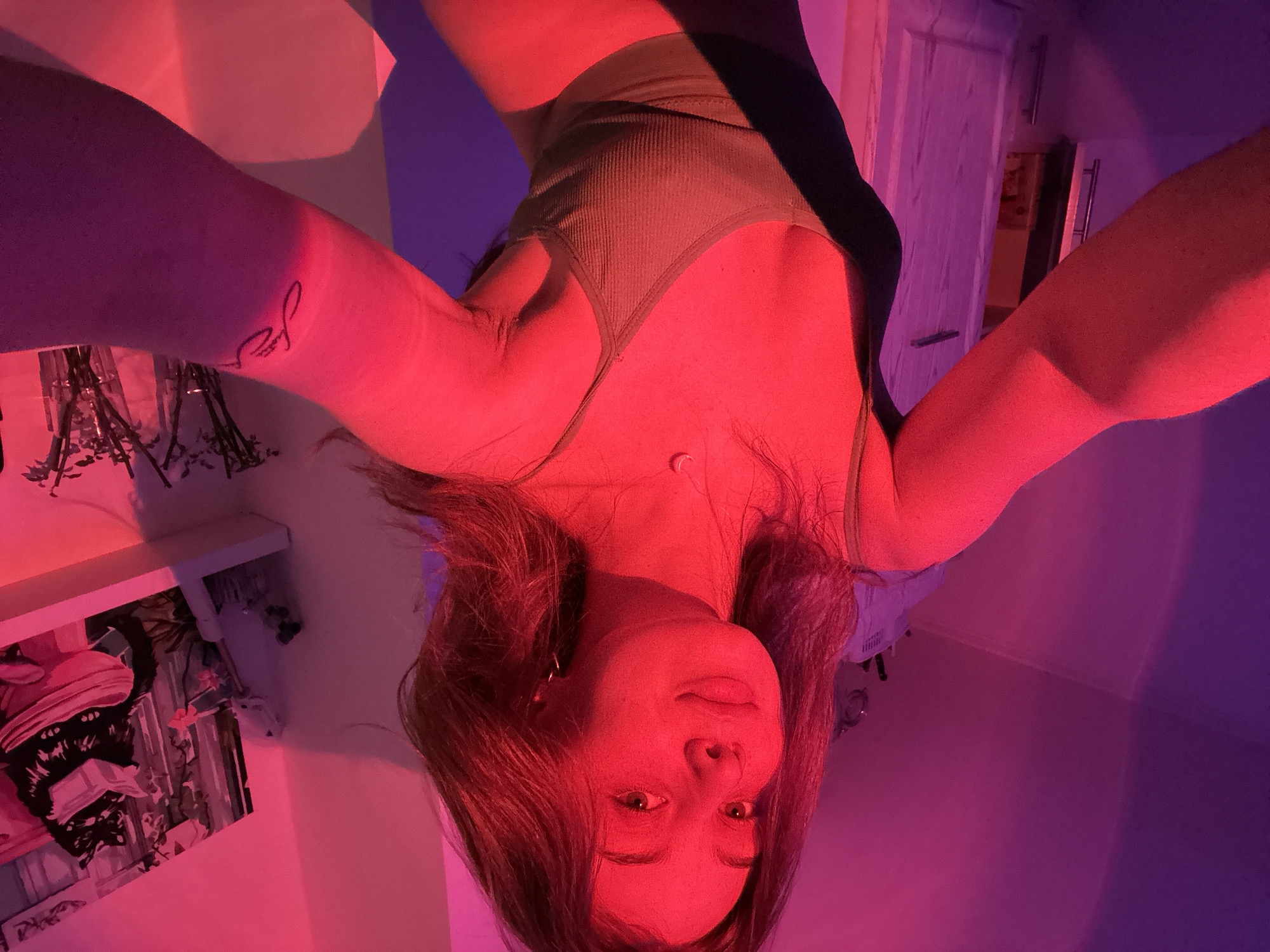 A selfie of a woman with long brown hair, wearing a green tank top, taken from an upside-down angle. The lighting appears to be a combination of pink and purple hues, casting a red tint over her face and skin. A tattoo is visible on her right arm. In the background, blurred objects suggest a room interior, including a door and shelving with items. The location is indeterminable.