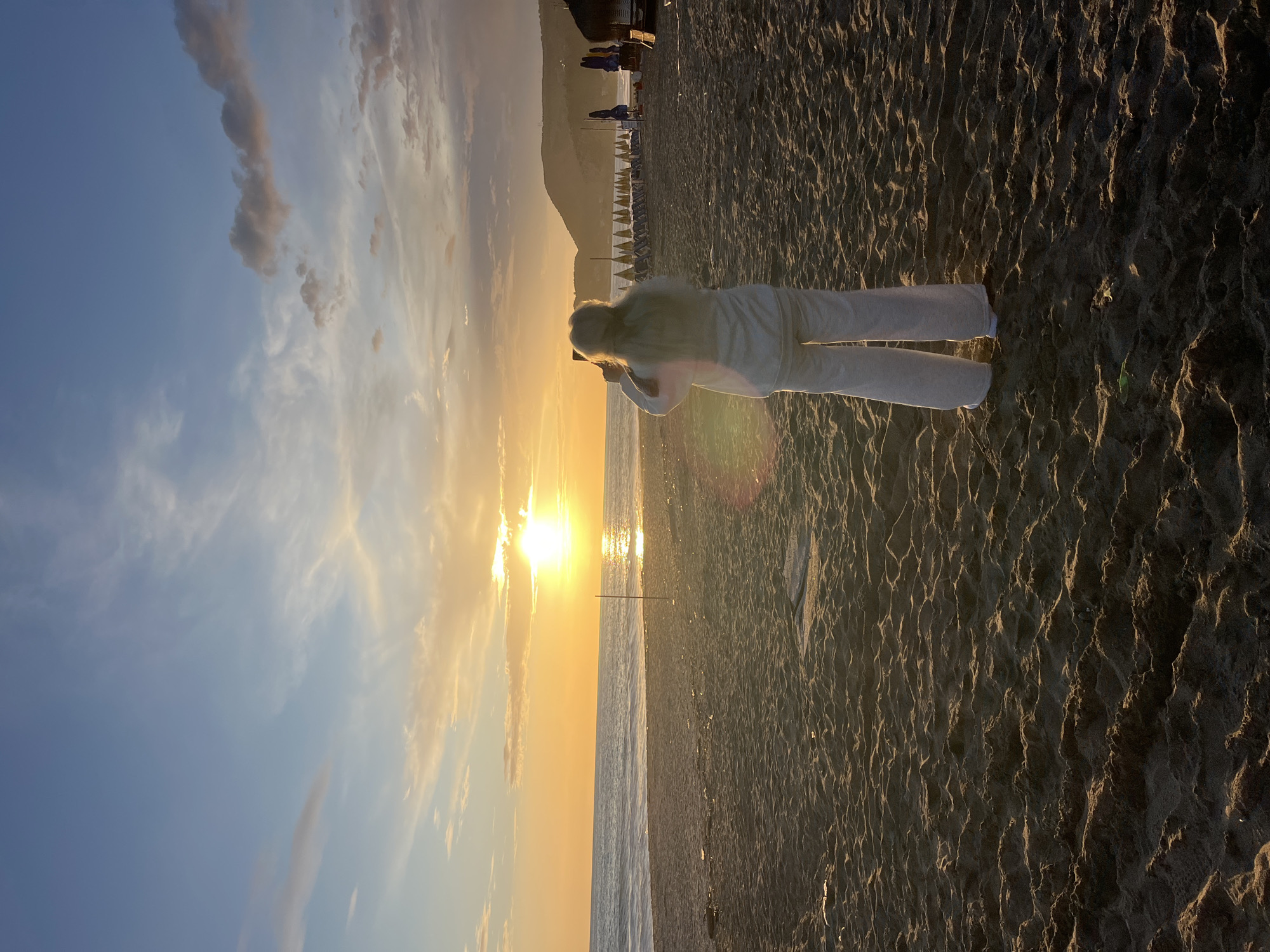 A person in light-colored clothing stands on a sandy beach, possibly in Alanya, Turkey, given the beach umbrellas in the background and the coastal setting. The individual appears to be taking a picture toward the setting sun, which is positioned directly in front of them, casting a strong glare and reflecting on the water. The beach has a texture of sand, while the sky displays a gradient of colors, from blue to orange, suggesting either sunrise or sunset. The horizon is defined by the sea. In the distance, there appear to be beach chairs and umbrellas, and a land formation is visible.