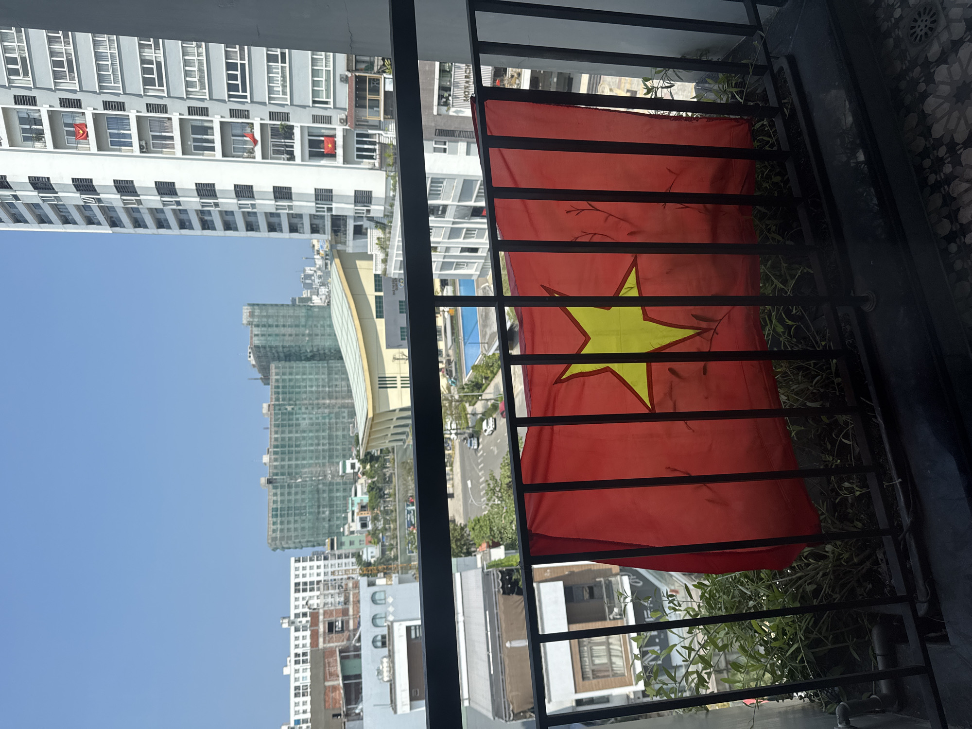 A medium-shot photograph taken at an oblique angle depicts the cityscape of Đà Nẵng, Vietnam, viewed from a balcony. The focal point is a Vietnamese flag hanging on the balcony railing. The flag is predominantly red, featuring a centered yellow star. The railing, constructed from black metal bars, obscures parts of the cityscape.
In the background, tall buildings dominate the skyline. Several structures appear to be under construction. The sky is a clear, light blue. Below the buildings, a street with vehicles is visible, lined with trees. Some greenery also grows from the bottom of the balcony. The scene suggests an urban environment with ongoing development.