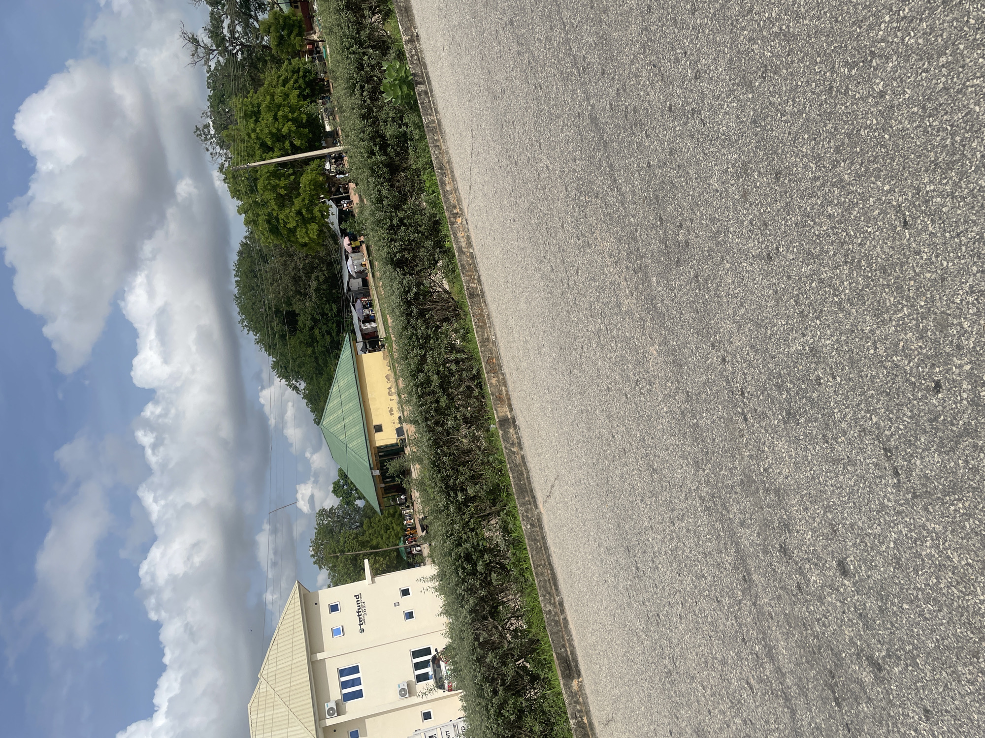 This image shows a view of an area in Jalingo, Nigeria, from the perspective of a street. In the foreground, a paved road is visible with a concrete curb and a dense green hedge bordering it. Beyond the hedge, there are buildings and trees. On the left, a light-colored, multi-story building with blue-framed windows and air conditioning units is seen. A sign on this building reads "TETFUND ANNUAL SUBVENTION 2024". To the right of this building, a yellow building with a green, sloped roof is present. In front of the yellow building, there are several stalls with umbrellas, suggesting a market or outdoor service area. People are visible in this area, as are trees and utility poles. The sky above is partly cloudy with large, white clouds against a blue sky.