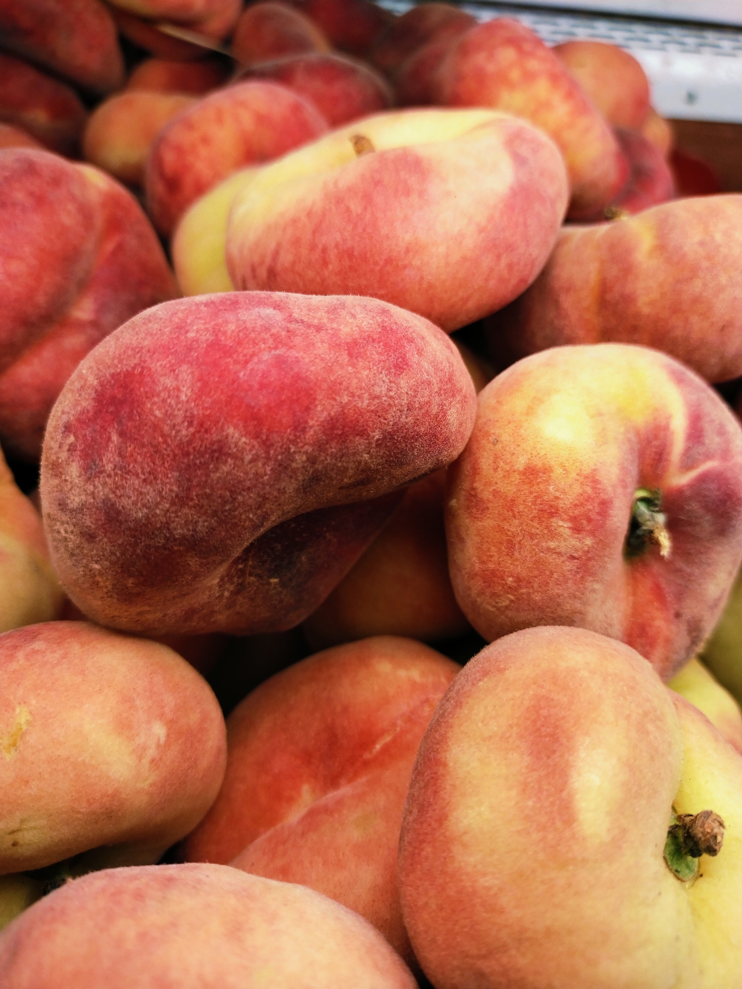 A close-up, overhead shot shows a pile of ripe Saturn peaches. The peaches are a mix of light pink, yellow, and red, with fuzzy skin and a slight dimple where the stem was attached. They are arranged in a tight, overflowing cluster, suggesting they are likely for sale at a market or produce stand. The background is blurred, focusing all attention on the textured surface of the fruit. There are no people or any other objects visible. The scene is likely taking place in Horishni Plavni, Ukraine, as this type of close-up fruit photography is common in market stalls there.
