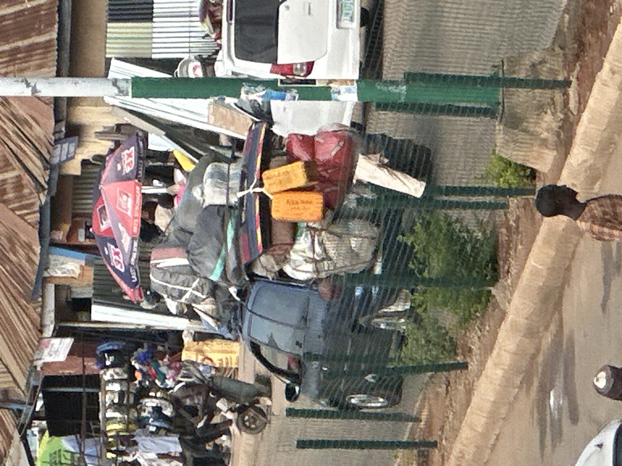 The image shows a busy street scene in Bukuru, Nigeria, likely a market or transport hub. A dark blue car is heavily loaded with luggage on its roof, including what appears to be several large bags and other bundled items. The car is parked on the side of the road, with a white van visible behind it. In the foreground, a dark green fence runs along the road. To the left of the car, a person on a motorcycle is visible, also appearing to be loaded with cargo. Market stalls with corrugated metal roofs and various goods are visible in the background, with umbrellas set up, one red and one blue, likely for shade. Several people are present in the scene, some seemingly engaged in activities related to trade or transport. The overall impression is one of a vibrant and active urban environment.