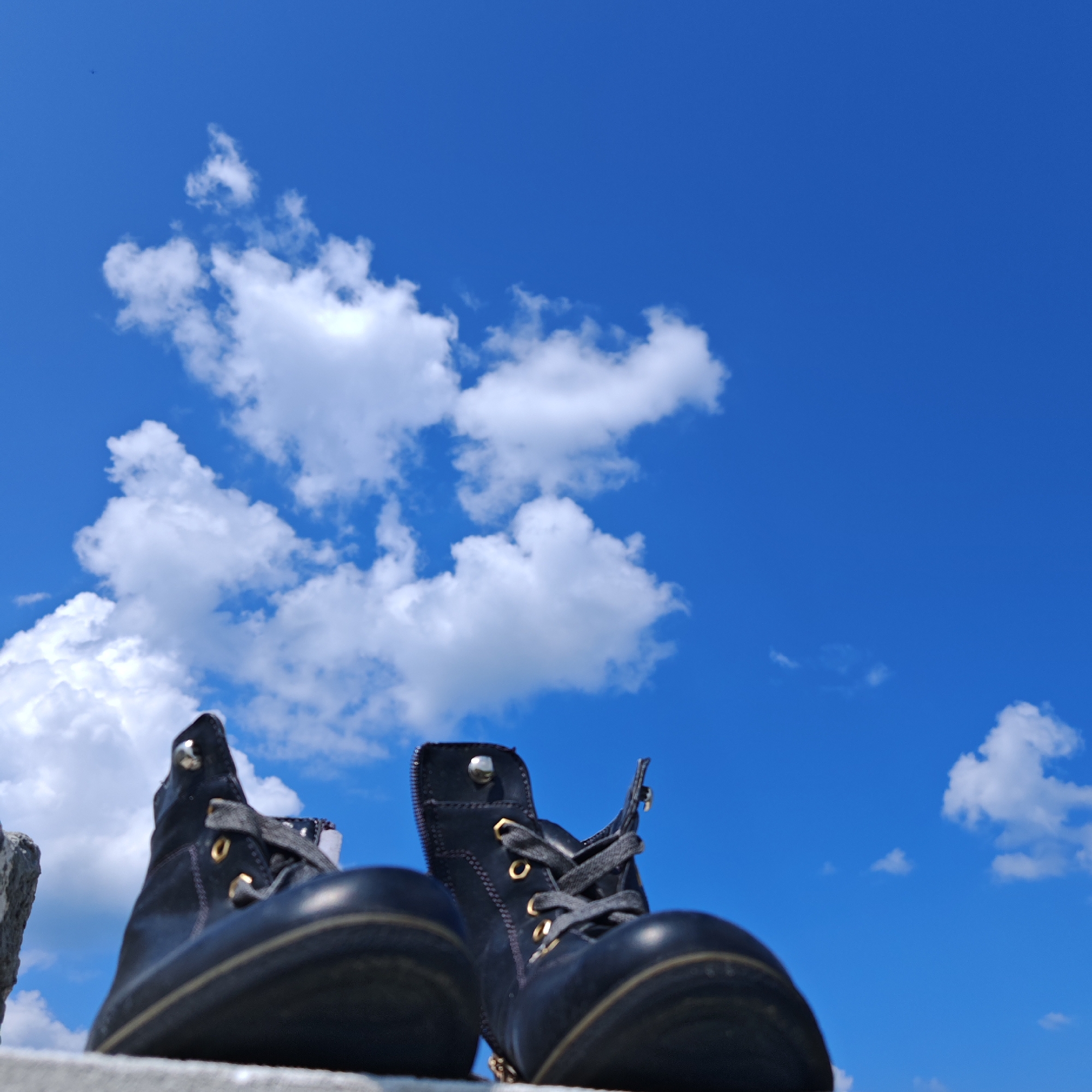 A low-angle, wide shot captures two black, lace-up boots positioned on a textured, light-colored surface against a vibrant, cloud-dotted blue sky. The boots are dark and appear to be made of leather, with gold-colored eyelets and dark grey laces. One boot is positioned slightly to the left and closer to the foreground, with its sole and side visible. The other boot is to the right, seen from a more frontal perspective, revealing its lacing and the top of its shaft. A small, dark object, possibly a bird, is faintly visible in the upper left quadrant of the sky. The scene suggests an outdoor setting, possibly a rooftop or elevated walkway, with a clear sky indicative of fair weather. There are no people visible in the image. The specific location within the city of Kurgan, Russia, cannot be determined from the visual information provided.