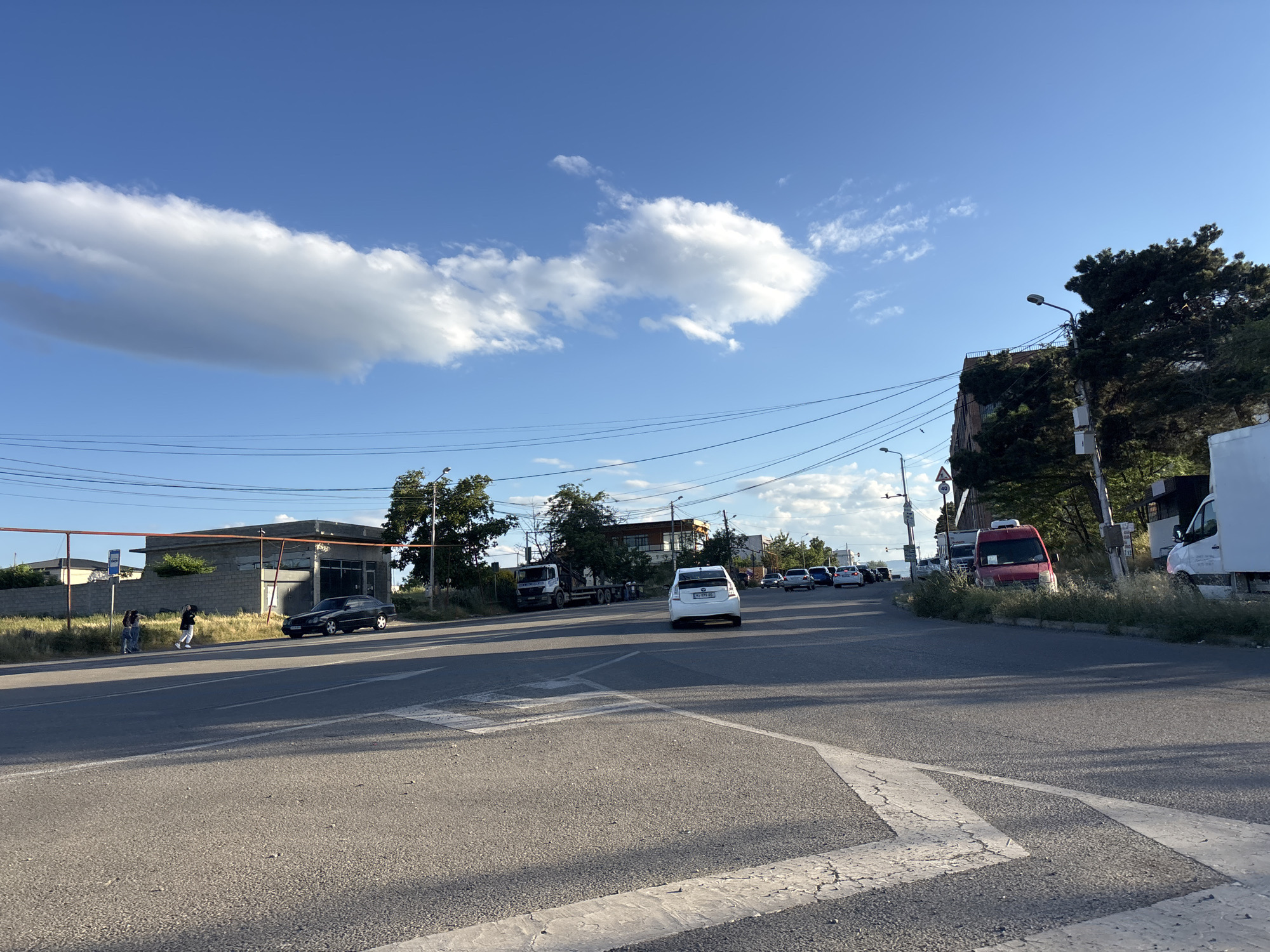 A street scene in Tbilisi, Georgia, captures a sunlit day with a bright blue sky dotted with fluffy white clouds. The perspective is from the road, looking towards an intersection or a slight curve in the road.
In the foreground, the asphalt road dominates, marked with white directional arrows. To the left, a low-lying building with a gray concrete facade is visible, with a few people standing near a bus stop sign. A black car is parked to the right of this building.
Further down the road, a white car is seen driving away from the camera, followed by several other vehicles. On the right side of the road, a red van and a white truck are parked, with lush green vegetation growing around them. Trees and utility poles line the roadside, with power lines stretching across the sky. A tall pine tree stands prominently on the right.
The scene appears to be set in the late afternoon or early evening, as indicated by the long shadows cast across the road and the warm, soft lighting. The weather is clear and pleasant, typical of a summer day. No specific event or strong emotion is discernible; it is a snapshot of everyday life on a suburban or semi-urban street. The overall impression is one of calm and ordinary activity.