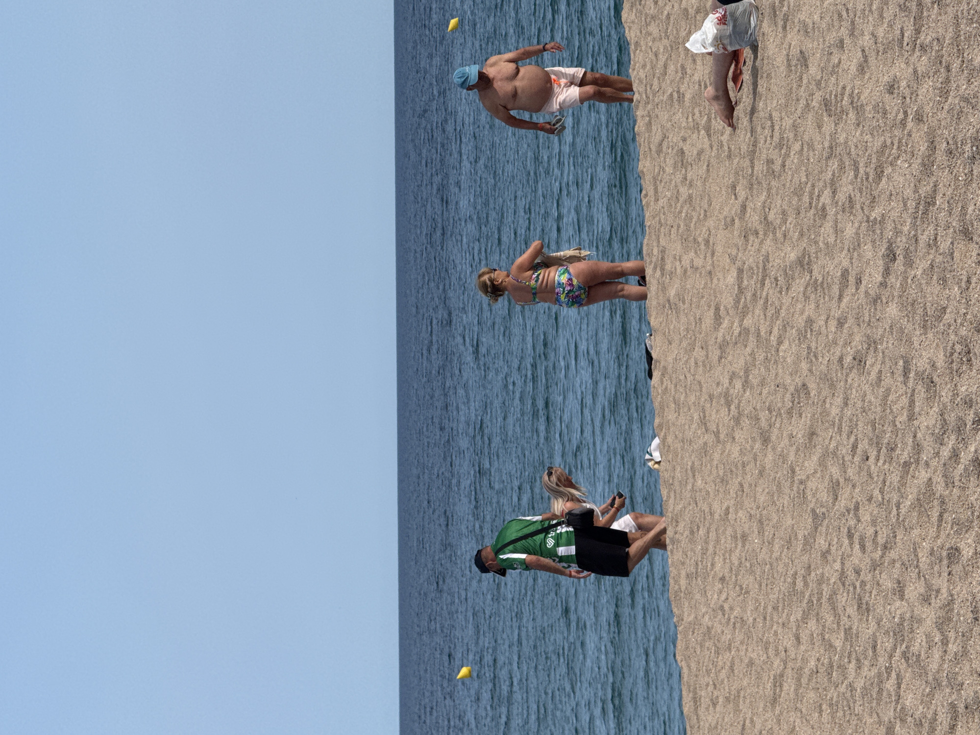 In Blanes, Spain, on a sandy beach, several people are observed near the water's edge. A man wearing a green and white striped shirt and black shorts, with a black bag slung over his shoulder, is walking towards the left. Beside him, a woman with blonde hair is bending down, looking at her phone. Further to the right, a woman in a floral bikini is holding a towel, facing left. A man in pink swim shorts and a blue hat is walking towards the right, holding a pair of flip-flops. In the background, the blue ocean stretches to the horizon under a clear sky, with yellow buoys visible in the water. Another person's legs are visible lying on the sand to the far right, partially obscured by a white plastic bag.