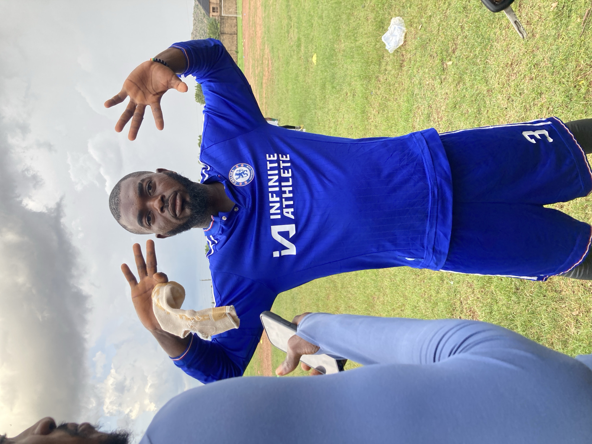 A low-angle, portrait-oriented shot depicts a man posing on a grassy sports field under an overcast sky in Jos, Nigeria. The main subject, a dark-skinned man with a beard, is wearing a blue long-sleeved athletic shirt and blue shorts. His shirt features the "Chelsea Football Club" logo on the left chest and the text "INFINITE ATHLETE" printed vertically below it in white. The number "31" is visible on his right short leg. He sports a dark beaded bracelet on his right wrist.
He is looking directly at the camera, making hand gestures: his right hand is extended towards the camera with fingers spread, while his left hand holds up two fingers (a peace sign) and clasps a light-colored, possibly bandaged or wrapped item.
In the foreground, partially visible, is another person's head and arm, holding a dark smartphone, suggesting they are the one taking the photograph. This person is wearing a light blue long-sleeved top.
The background shows a green grassy field with some white litter and a car key scattered on it. A faint yellow cone and a building structure can be seen in the distance. The sky is largely covered with grey clouds, indicating overcast weather during the daytime. The man's expression is neutral to slightly smiling, conveying a relaxed pose.