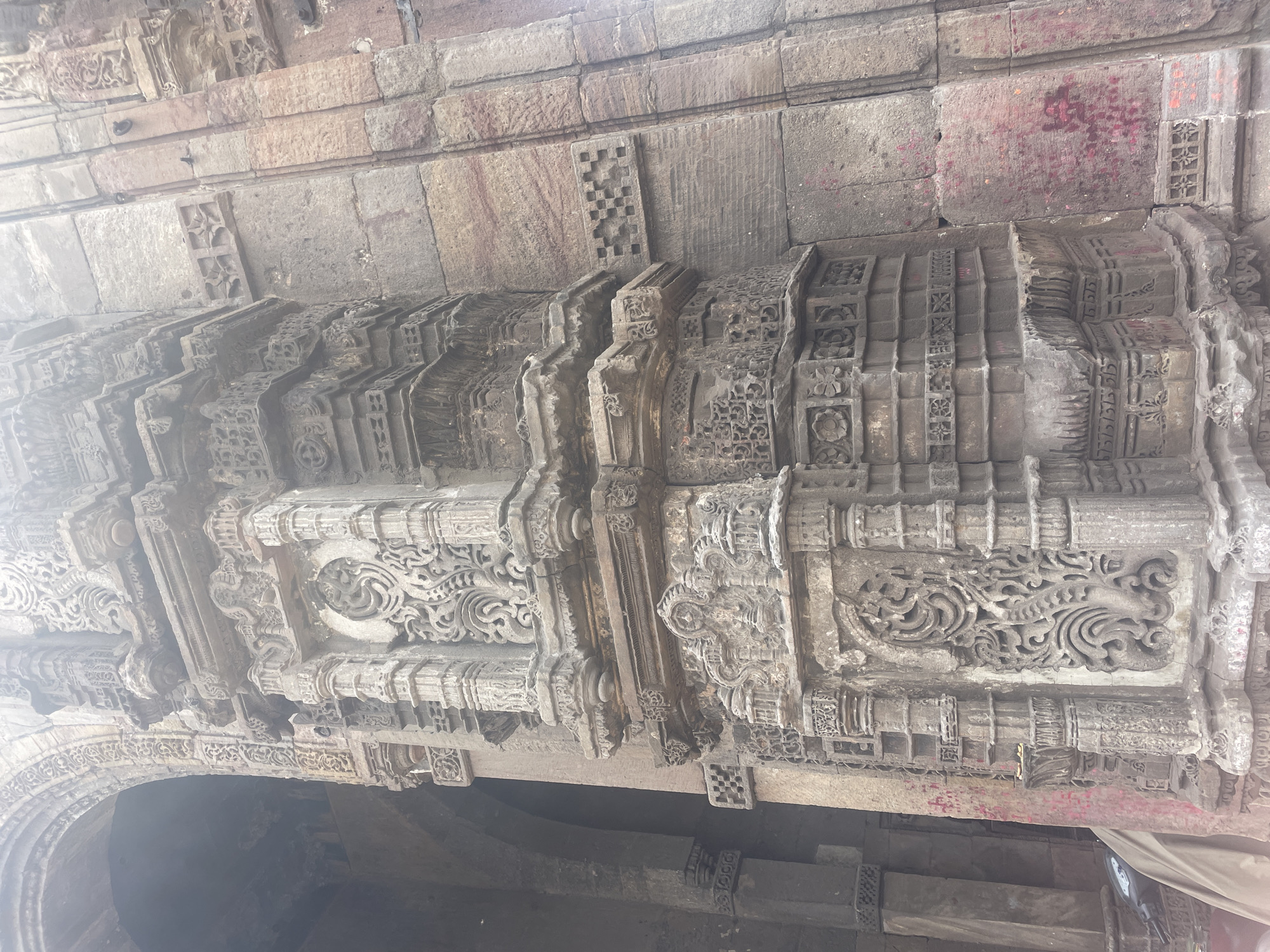 The image shows a close-up view of the intricately carved stone architecture of the Sidi Saiyyed Mosque in Ahmedabad, India. The focus is on a section of the mosque's facade, highlighting the detailed stonework. The stone is weathered and appears to be sandstone, with shades of grey and reddish-brown. The carvings include floral motifs, geometric patterns, and decorative columns. The sunlight creates a bright, hazy effect at the top of the image, suggesting it might be daytime. Part of an arched entrance or alcove is visible on the left, and a draped object, possibly a tarp covering something, is partially visible at the bottom left. The overall impression is one of ancient, artistic craftsmanship.