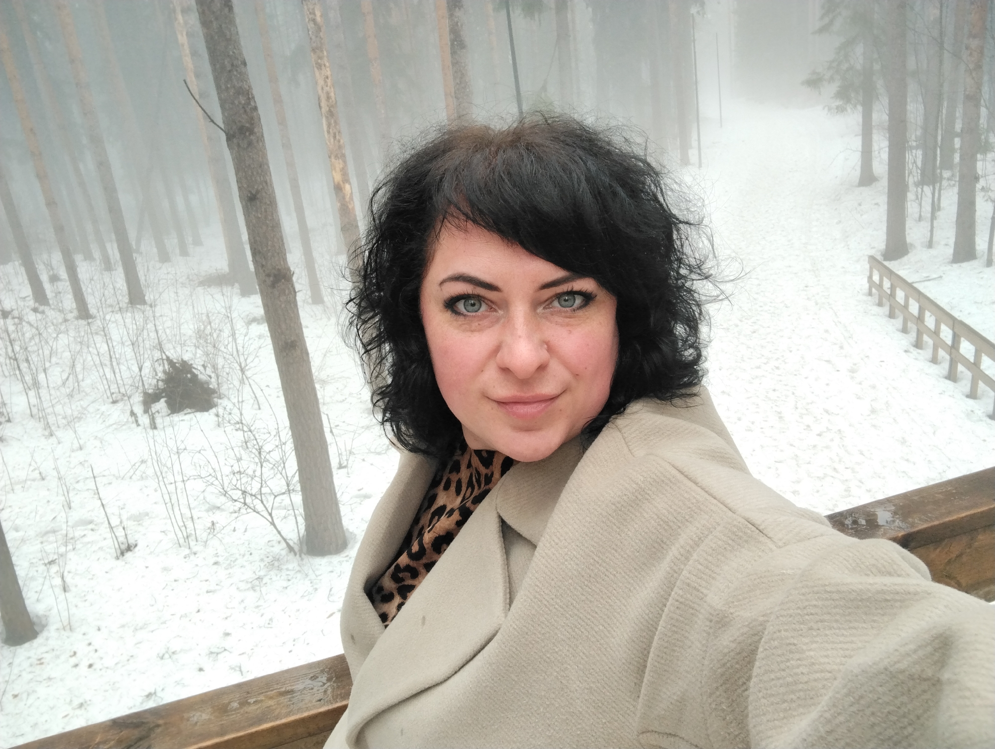 A woman with dark curly hair and light-colored eyes is taking a selfie outdoors. She is wearing a light beige coat over a garment with a leopard print pattern. The background shows a snowy landscape with tall, thin trees, indicating a wooded area. The weather appears to be foggy, obscuring details in the distance. The woman stands on a wooden structure with a railing, overlooking the snowy terrain. Based on the location of the image being taken in Votkinsk, Russia, it is likely that this is a forest or park in the region during winter.