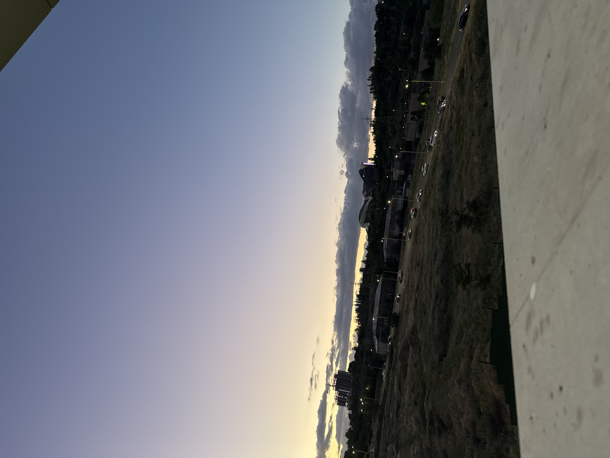 The image captures an expansive twilight or dawn scene from an elevated perspective, likely a balcony or high floor overlooking a landscape. The sky dominates the upper half, transitioning in color from a light, clear blue overhead to a warm, soft yellow and orange glow along the horizon. Several long, narrow clouds stretch horizontally across the sky near the horizon line.
In the middle ground, a wide expanse of dark, low-lying vegetation or dry grass is visible, traversed by a road. Numerous cars are present on the road, identifiable by their illuminated headlights and taillights, suggesting low light conditions. Streetlights dot the roadside, casting pools of light. Further in the distance, a darker line of trees or hills forms a silhouette against the fading sky. Scattered buildings, some with visible lights, and what appears to be a structure under construction with scaffolding are discernible in the distance. A power line pylon is also visible.
A textured, light-colored concrete or stone wall occupies the far right side of the foreground, indicating the barrier of the viewing platform. No visible text is present. The overall atmosphere is calm and transitional, characteristic of early morning or late evening. The scene depicts a semi-urban or suburban landscape, consistent with a view over Yerevan, Armenia.