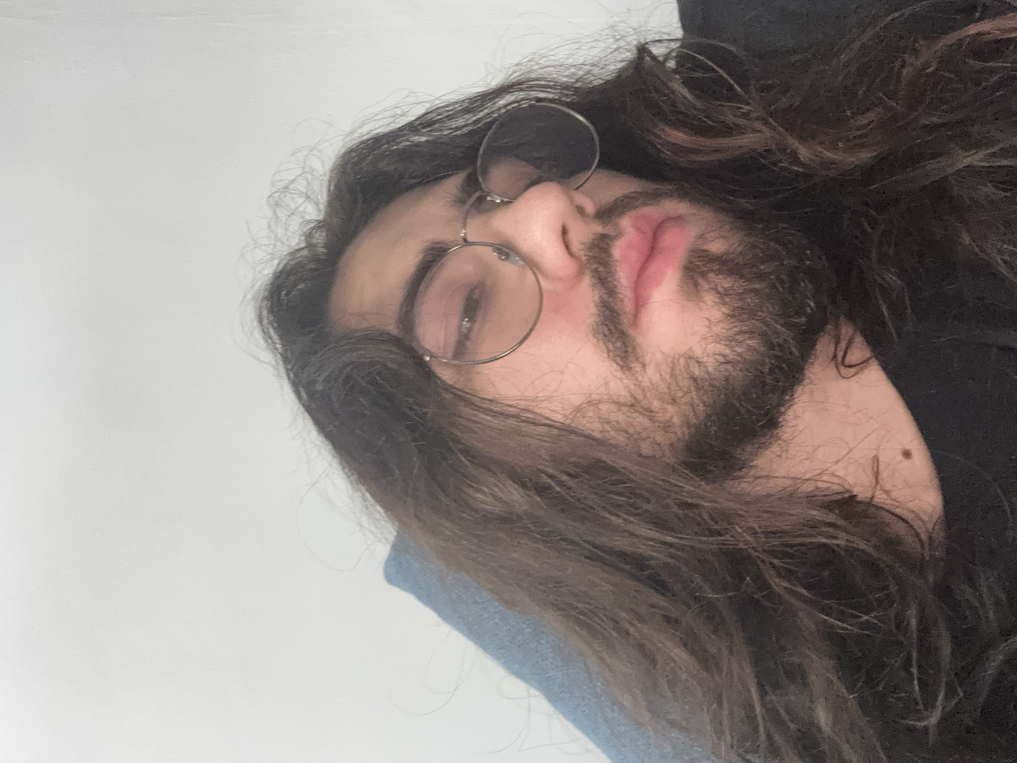 A man with long, dark, wavy hair and a beard wears round, wire-rimmed glasses and a black t-shirt. He is looking slightly down and to his right, with his lips slightly parted. The background is a plain white wall. The lighting suggests an indoor setting, possibly a home. No specific location within a city can be determined from the image.
