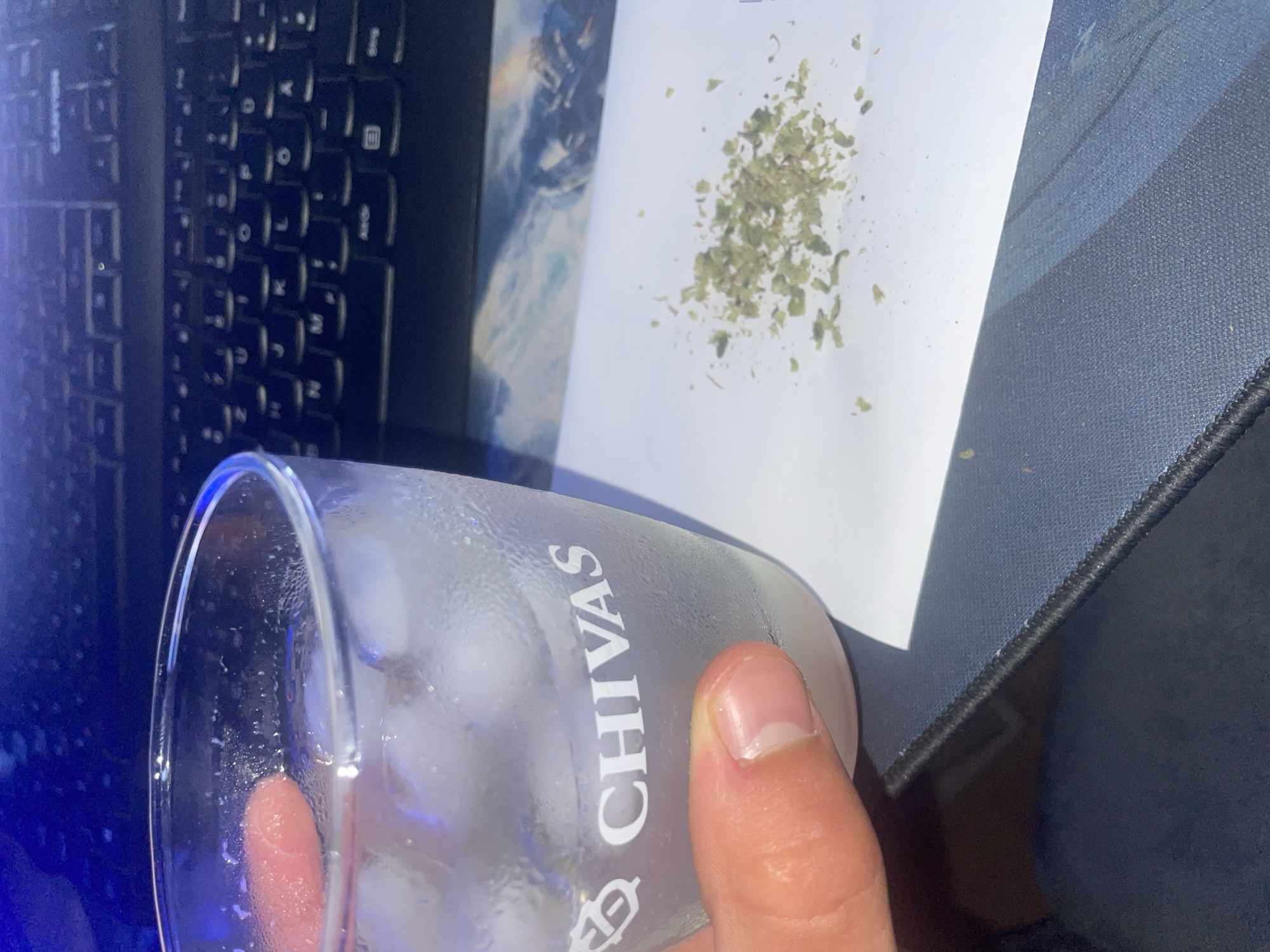 An overhead, close-up shot shows a person's hand holding a glass of ice water that has condensation on the outside. The glass has the Chivas logo on it. Next to the glass is a small pile of cannabis. In the background, a black keyboard is visible. The scene is not in a city.