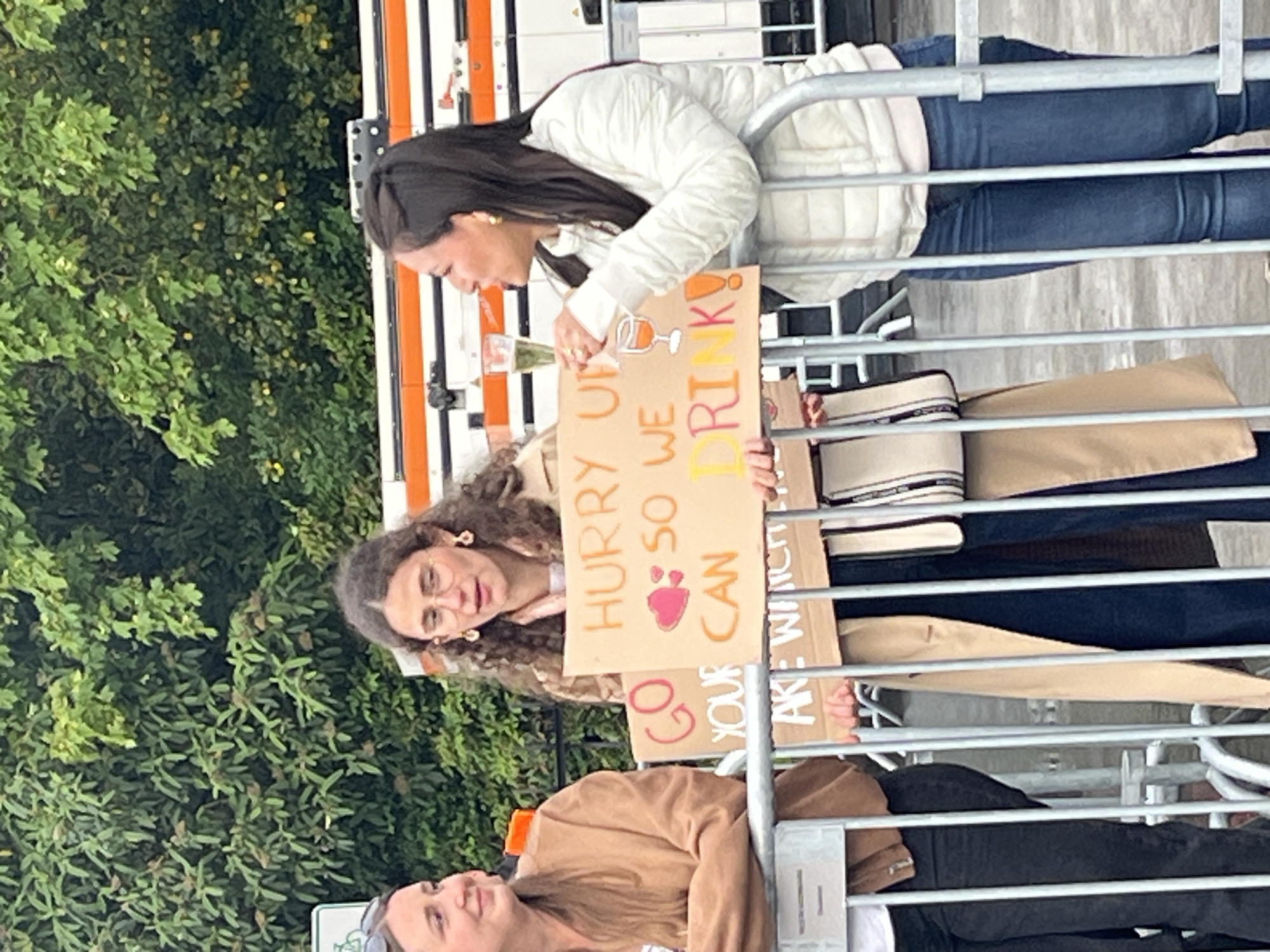 Three women stand behind a metal barrier, likely at an event or parade in Luxembourg City, Luxembourg. One woman holds a sign that reads "Hurry up so we can..." with a drawing of a wine glass and "drink". Another sign reads "Go your way, we are watching". The women are dressed casually in sweaters and jeans. The environment is a mix of greenery and urban architecture.