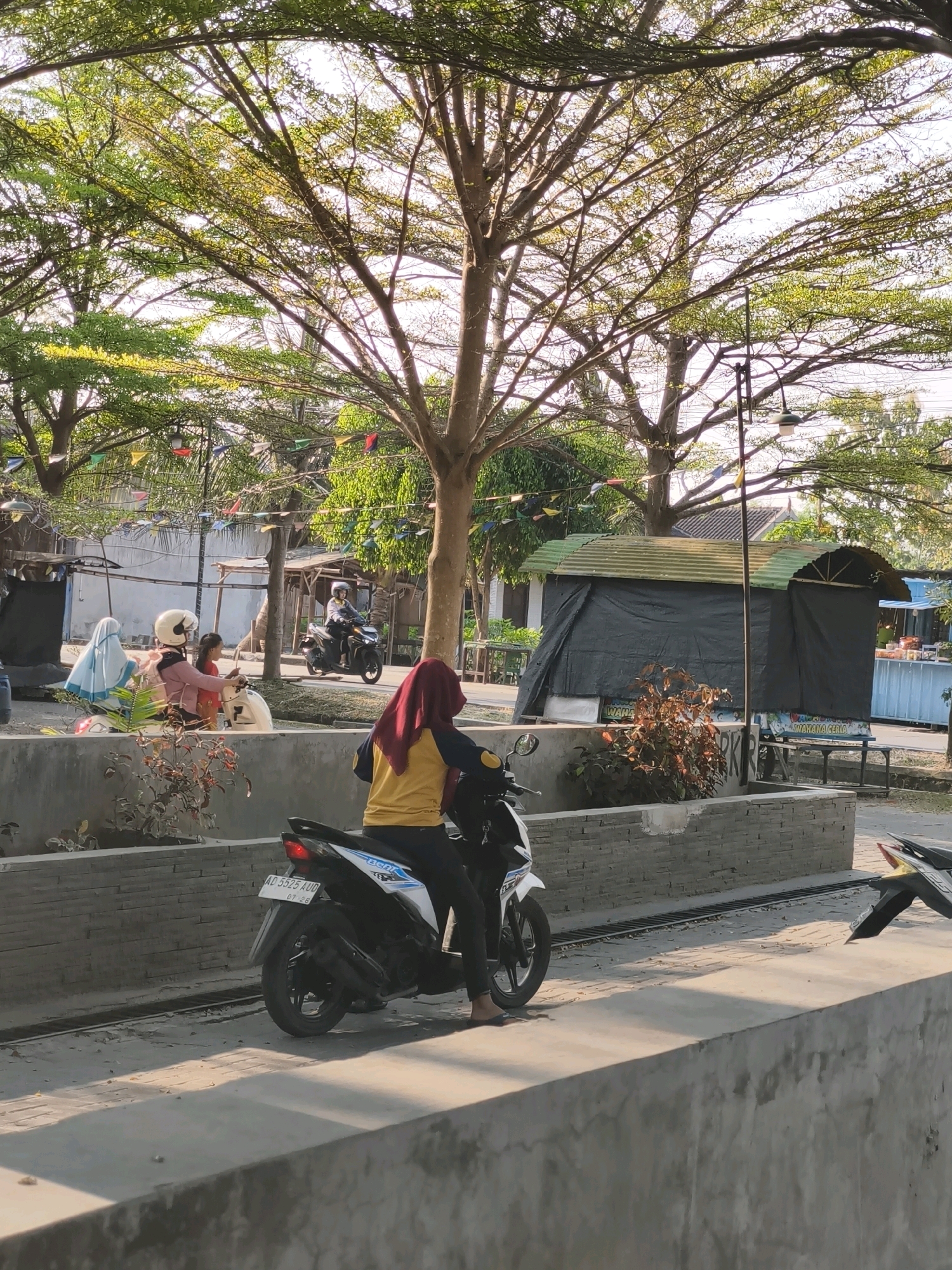 In Desa Dibal, Indonesia, a woman wearing a red hijab and a yellow and blue shirt is riding a white motorcycle. She is positioned on the right side of the frame, with the motorcycle facing towards the left. In the background, another motorcycle carrying two people is visible, moving from left to right. The scene is set outdoors, with a large tree with lush green foliage dominating the upper portion of the image. Colorful triangular flags are strung between trees, suggesting a festive atmosphere or a market setting. Several structures, including a dark-colored stall with a corrugated metal roof, are present in the mid-ground. A concrete barrier runs across the lower part of the image, with a drain grate visible in front of it. The lighting indicates it is daytime, with sunlight casting shadows on the ground.