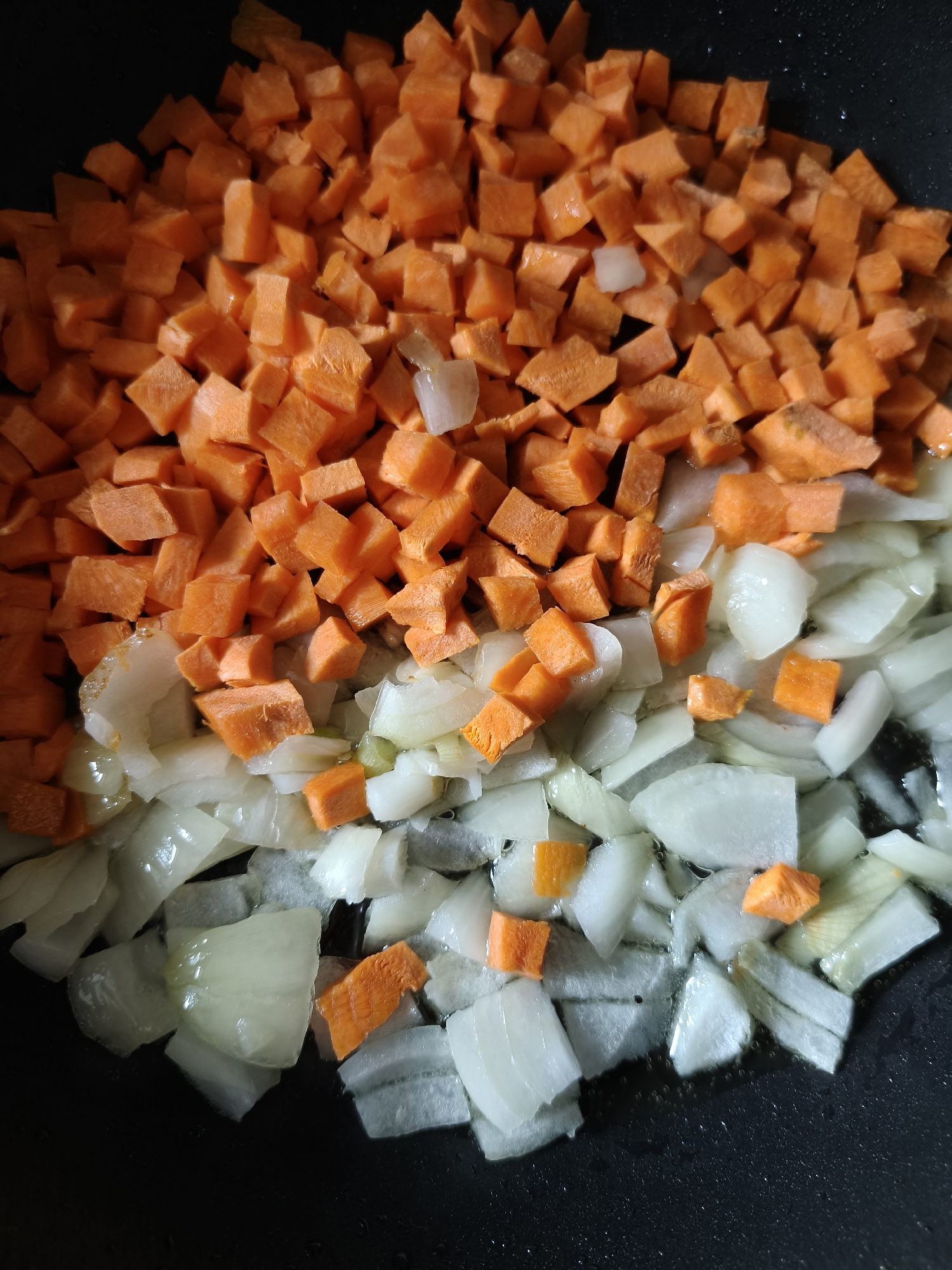The image shows diced carrots and onions in a black pan. The carrots are bright orange and cut into small cubes, occupying the top portion of the pan. The onions are translucent white and appear to be sautéed or cooking with some moisture in the bottom of the pan. The location is not discernible from the image.