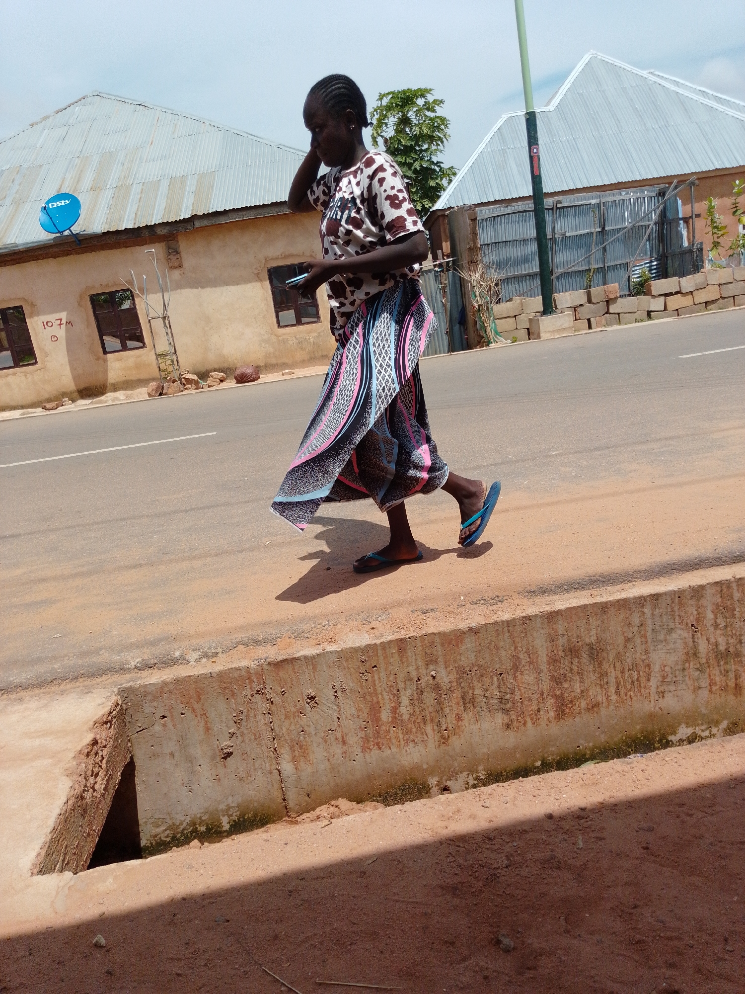 A woman is walking along a dirt path next to a road. The woman is wearing a white shirt with brown spots and a long, colorful skirt. She is holding a mobile phone in her right hand and appears to be talking on it, with her left hand near her ear. She is wearing blue flip-flops. In the background, there are buildings with corrugated metal roofs and some vegetation. The weather appears to be cloudy. The image is taken from a slightly low angle, looking up at the woman. The overall scene suggests an outdoor urban or semi-urban environment.