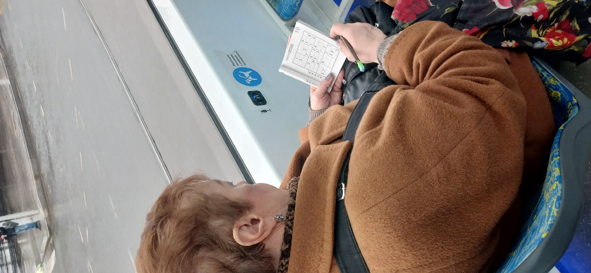 A woman is seated, facing away from the camera, while riding in a public transportation vehicle. She is engrossed in a Sudoku puzzle, holding the paper with one hand and a pen in the other. The woman has short, reddish-brown hair and is wearing a brown coat. A black bag is slung over her shoulder, and a floral patterned item rests beside her on the seat. The interior of the vehicle has a white wall with a sign and an electronic device. Outside, a sidewalk and building exteriors are visible through the window. The location is Unknown, Unknown.