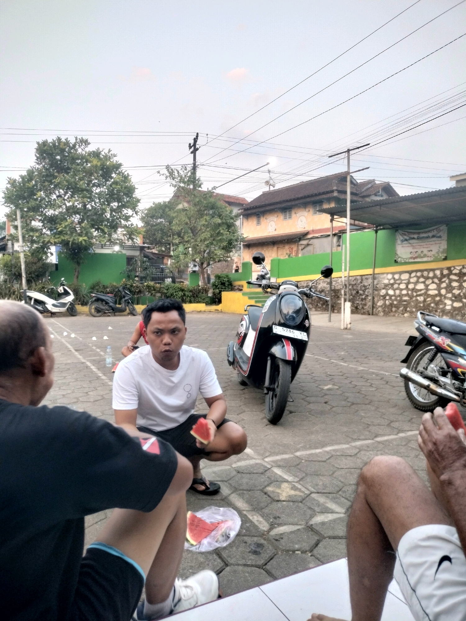 This is a photograph taken outdoors, likely in an urban or suburban setting in Boja, Indonesia. In the foreground, three individuals are partially visible. One person, on the left, is mostly obscured but appears to be wearing a black t-shirt. Another person, in the center, is a man wearing a white t-shirt and black shorts, crouching and holding a piece of watermelon. He is looking directly at the camera. To the right, another person is visible from the waist down, wearing white shorts and holding a slice of watermelon.
The background features several elements indicative of a developed area. There are two motorcycles parked on a paved surface. One is a black scooter with red and white accents. The other is a more traditional-looking motorcycle. Beyond the vehicles, there are trees, buildings with tiled roofs, and power lines crisscrossing the sky. A green wall and a stone wall are also visible. The overall impression is that of a casual gathering in a public space.