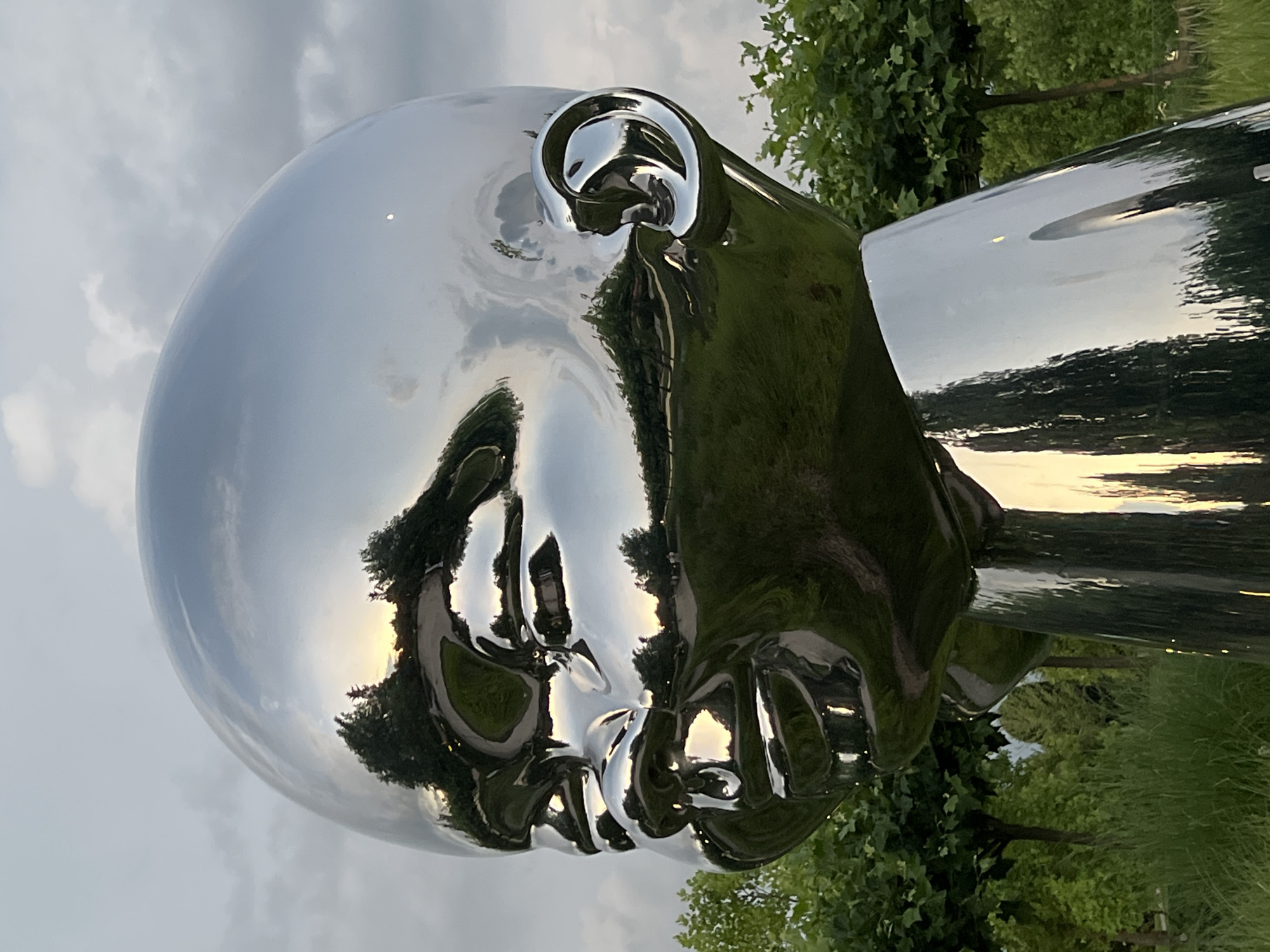 The image shows a close-up of a large, silver, reflective sculpture of a baby's head and neck. The sculpture is located outdoors in a park-like setting, with lush green grass and trees visible in the background. The sky above is overcast with some hints of sunlight breaking through. The reflective surface of the sculpture mirrors the surrounding environment, including trees, clouds, and a path. The sculpture is positioned in a way that it faces towards the left of the frame. The sculpture appears to be part of the Krasnodar Monument to the First Teacher, located in Krasnodar, Russia, and is a popular tourist attraction.