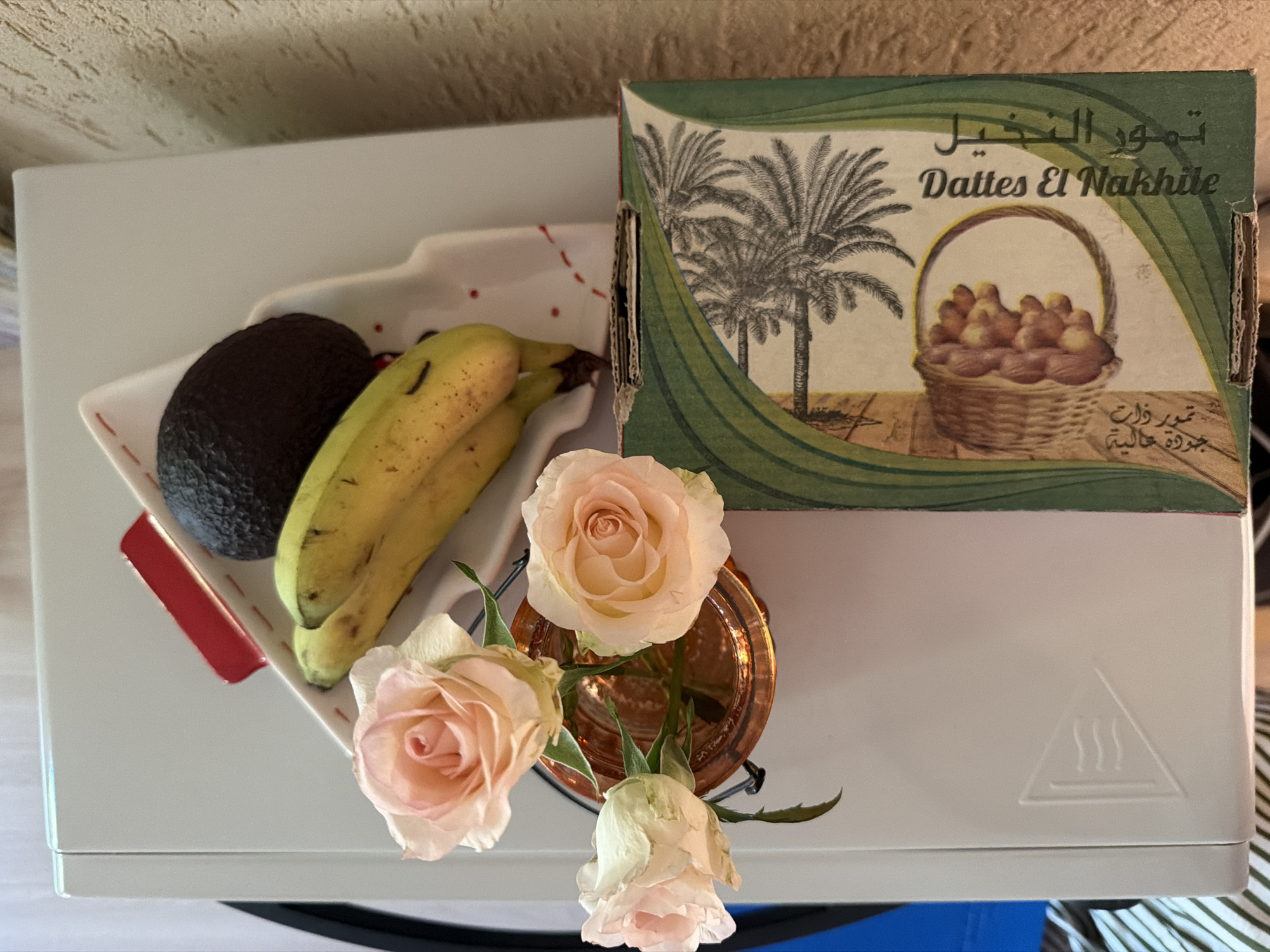 A still life arrangement is presented on a light-colored surface, possibly a piece of furniture. In the foreground, a small copper-colored vase holds three light pink roses. To the right and slightly behind the roses, a cardboard box with a green border features an illustration of palm trees and dates, with text in Arabic and English that reads "Dattes El Nakhile." Resting on a white dish with red stitching and red dots, a dark-skinned avocado and two yellow bananas are visible. The image has a shallow depth of field, with the roses and the box in sharper focus than the background. There are no people visible in the image. The location is not specified in the image.