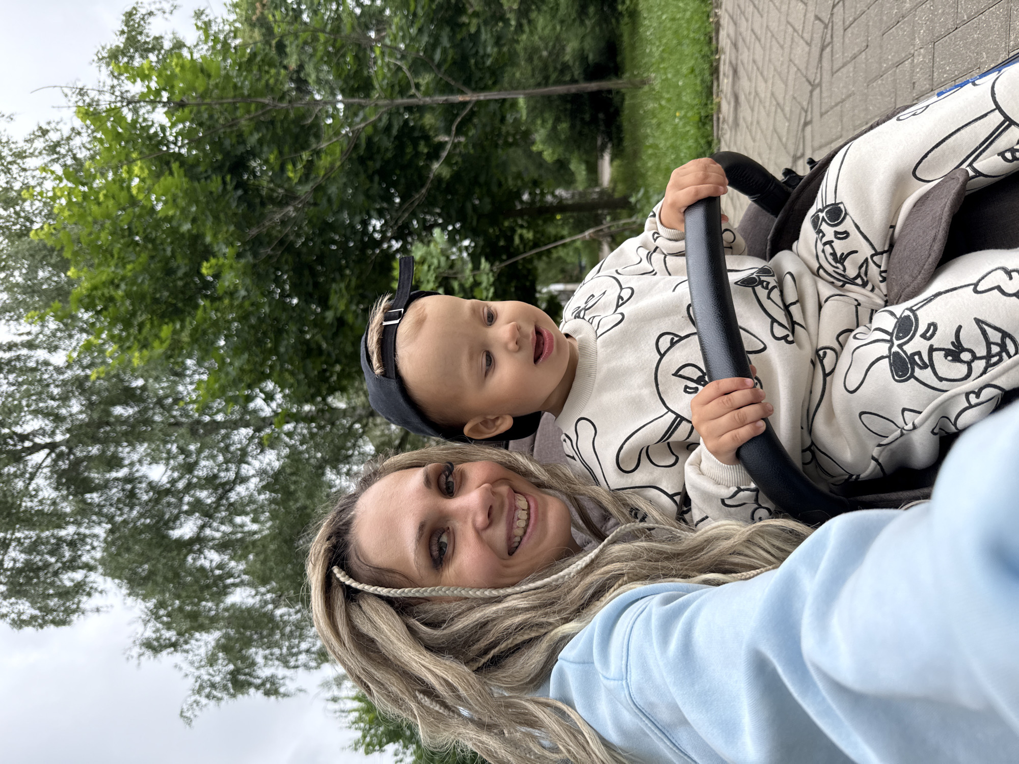 A woman with blonde dreadlocks and a light blue hoodie smiles at the camera, her arm extended to take a selfie. Beside her, a toddler wearing a black baseball cap and a white tracksuit with cartoon bunny prints looks to the side, grasping the handle of a stroller. They are positioned in a park, with lush green trees and a cloudy sky forming the background. The ground is paved with light gray bricks. The lighting is soft, suggesting an overcast day.