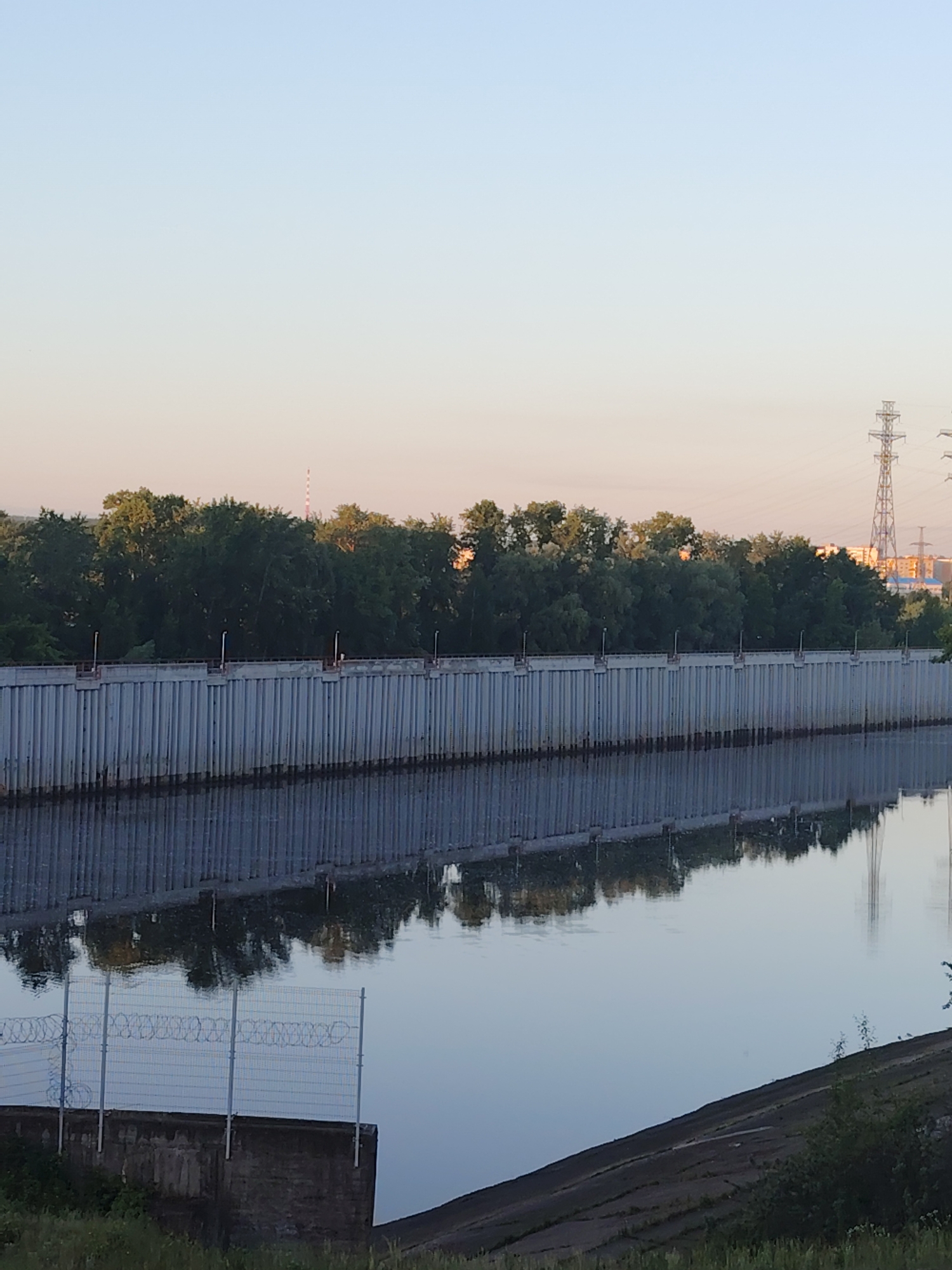 The image depicts a serene riverfront scene at dawn in Chaikovsky, Russia. A tall, ribbed concrete wall runs along the water's edge, topped by a row of evenly spaced metal posts with small lights. Behind the wall, a dense line of green trees forms a natural barrier, with a few distant buildings and a tall power pylon visible through the foliage. The calm river mirrors the wall and trees, creating a symmetrical reflection. In the foreground, a section of concrete embankment is visible with a wire fence topped with barbed wire, suggesting a security measure. The sky transitions from a pale pink near the horizon to a soft blue overhead. The overall impression is one of quiet industrial infrastructure integrated with natural elements.
