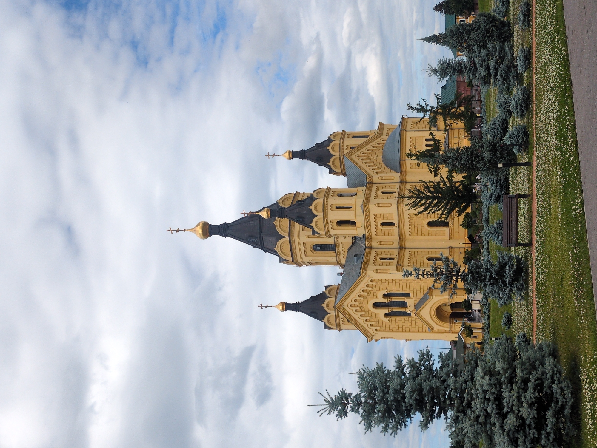 The image depicts a sunny day at the Alexander Nevsky Cathedral in Nizhny Novgorod, Russia. The cathedral, a prominent landmark of the city, is situated on a hill overlooking the Volga River. The cathedral is a large, ornate structure with multiple golden domes and intricate architectural details. In the foreground, lush green grass is dotted with small white flowers, suggesting a park-like setting. Several coniferous trees, varying in size and shade of green, are scattered throughout the scene. A wooden bench is placed on a paved path, inviting visitors to rest and admire the view. The sky is a mix of blue and white clouds, indicating fair weather. The overall impression is one of peace and tranquility, with the majestic cathedral serving as the centerpiece of the landscape.