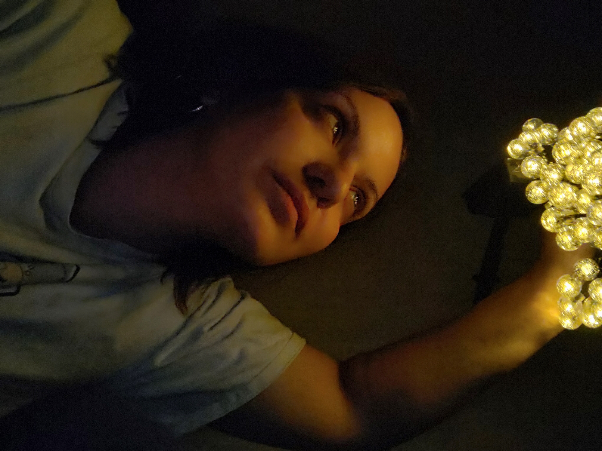 A woman is lying down in a dimly lit area, possibly indoors. She is wearing a light-colored t-shirt and holding a string of fairy lights in her hand. Her face is illuminated by the lights, while the background is mostly dark. The location is not discernible as Марковский, Russia, based on the image.