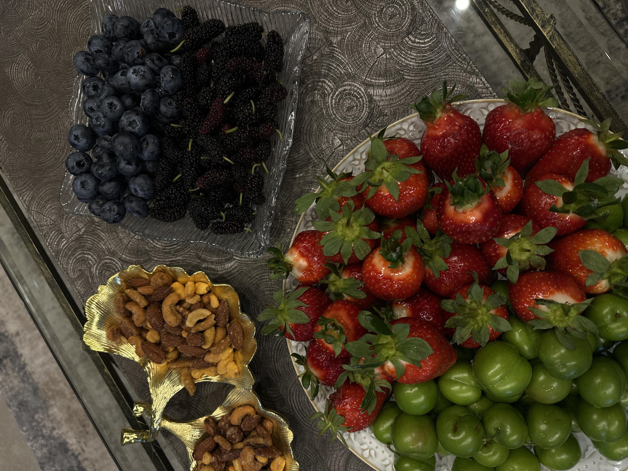 The image displays a spread of fresh fruits and nuts arranged on a textured surface, likely a glass tabletop. Located in Yerevan, Armenia, the scene presents an assortment of foods.
In the upper left corner, a clear glass dish holds blueberries and mulberries, each occupying half the dish. Adjacent to it, a white plate with gold trim contains a large pile of strawberries, under which is a smaller pile of green plums. Below the berries, a gold-colored dish shaped like leaves contains a mixture of nuts.
The lighting in the image is bright and appears to be artificial, given the shadows and highlights on the food items. The surface beneath the food items is textured with a pattern that resembles wood grain or concentric circles. The scene lacks people.