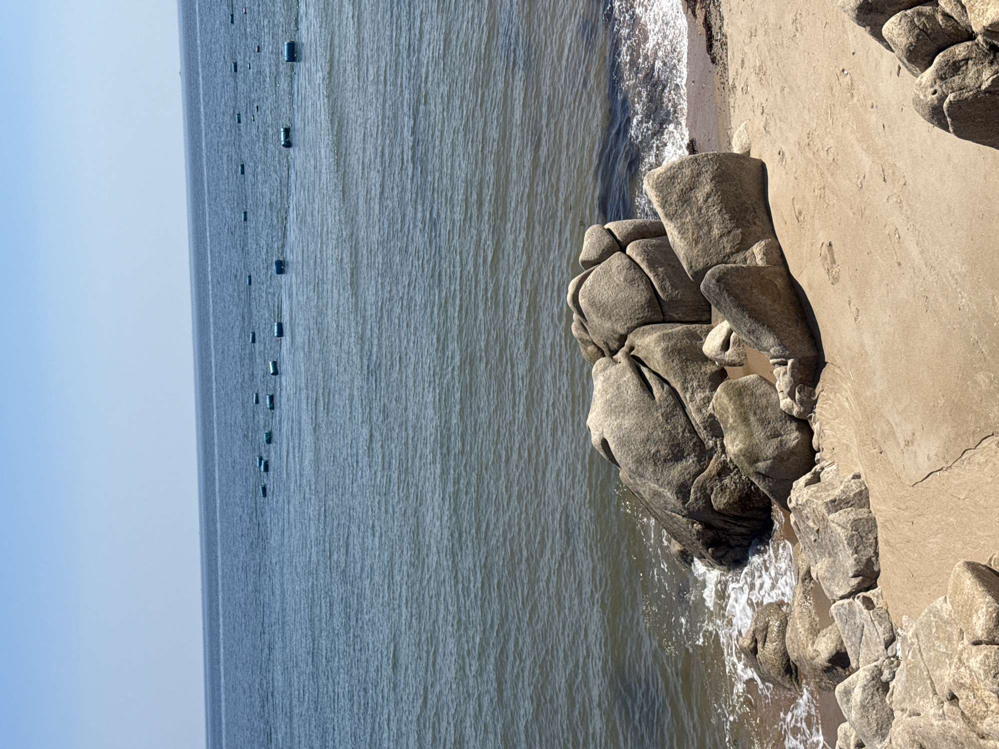 This image captures a tranquil scene at a beach in Qinhuangdao, China, on a clear, sunny day. The vast expanse of the blue sea stretches to the horizon, dotted with numerous small, blue buoys, suggesting aquaculture or navigational markers. The gentle waves lap onto the sandy shore, where large, weathered rocks are scattered. In the foreground, a cluster of these rocks, smoothed by time and tide, sits prominently on the sand. The sandy beach itself shows subtle imprints, possibly from footprints or the movement of the tide. The overall atmosphere is one of peaceful solitude, with no people visible in the frame.