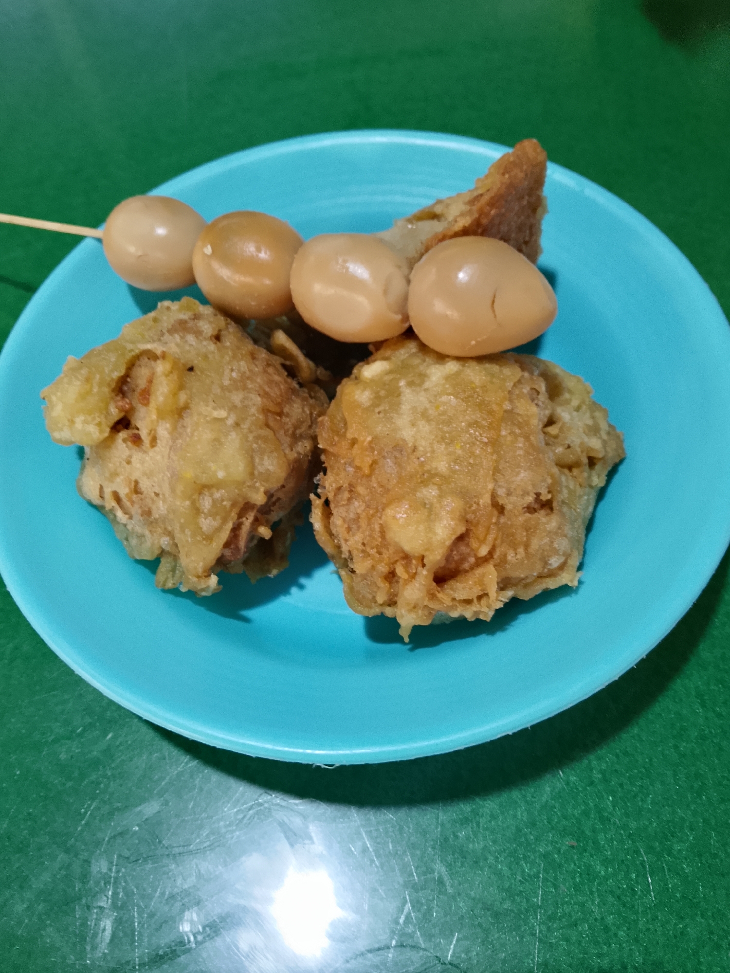 A close-up, overhead shot shows a bright teal plate filled with Indonesian street food. Two pieces of fried, golden-brown fritters are prominently featured, with a skewer of three hard-boiled quail eggs and a piece of fried bread positioned behind them. The food is set against a green, textured surface, likely a table. The lighting suggests an indoor setting. The image is of a prepared meal, likely in a casual dining environment such as a warung in Balun, Indonesia.