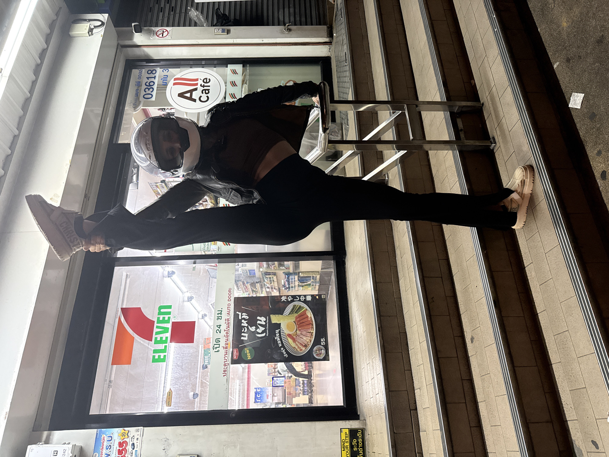 A person wearing a black leather jacket, dark top, black pants, a white helmet, and tan slip-on sneakers performs a high leg split with their right leg extended upwards. They are holding onto a metal railing for support. The person is standing on a set of stone steps in front of a 7-Eleven convenience store. The store's windows display the 7-Eleven logo, "All Cafe" signage, and promotional posters for food items. The surrounding environment appears to be an urban street at night, with artificial lighting illuminating the scene.