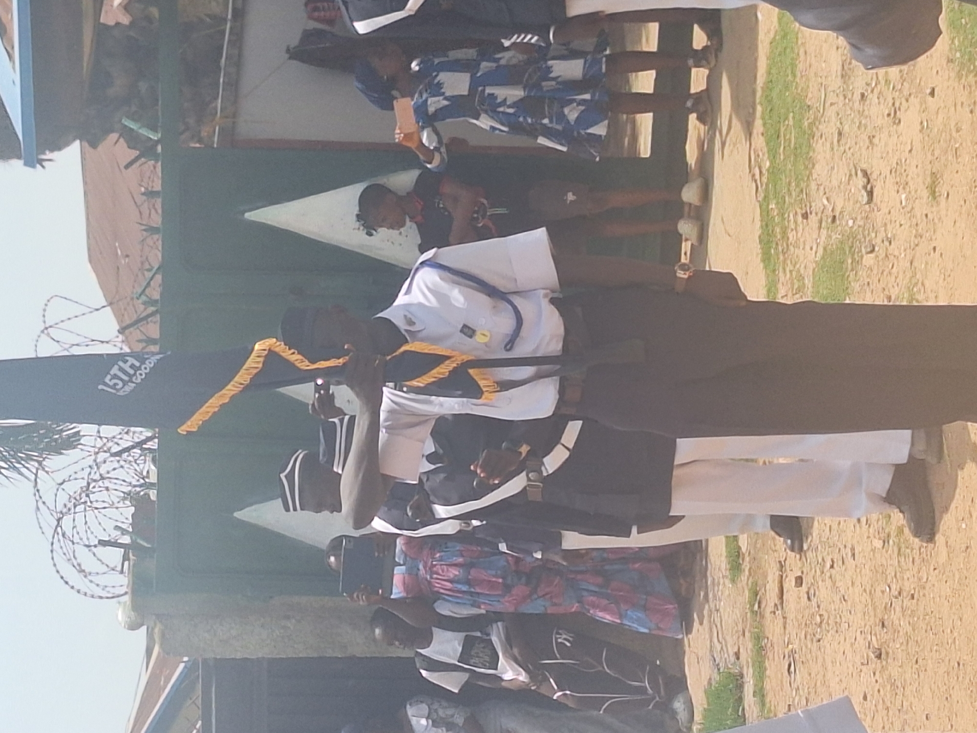 The image captures a scene in Sabon Tasha, Nigeria. A group of uniformed individuals is standing outdoors, near a building or structure that appears to be a gate or entrance, secured with barbed wire. One individual is holding a flag or banner, while others are taking pictures with their phones. The surrounding environment consists of a dirt or gravel surface. Civilians stand nearby, possibly observing the group.