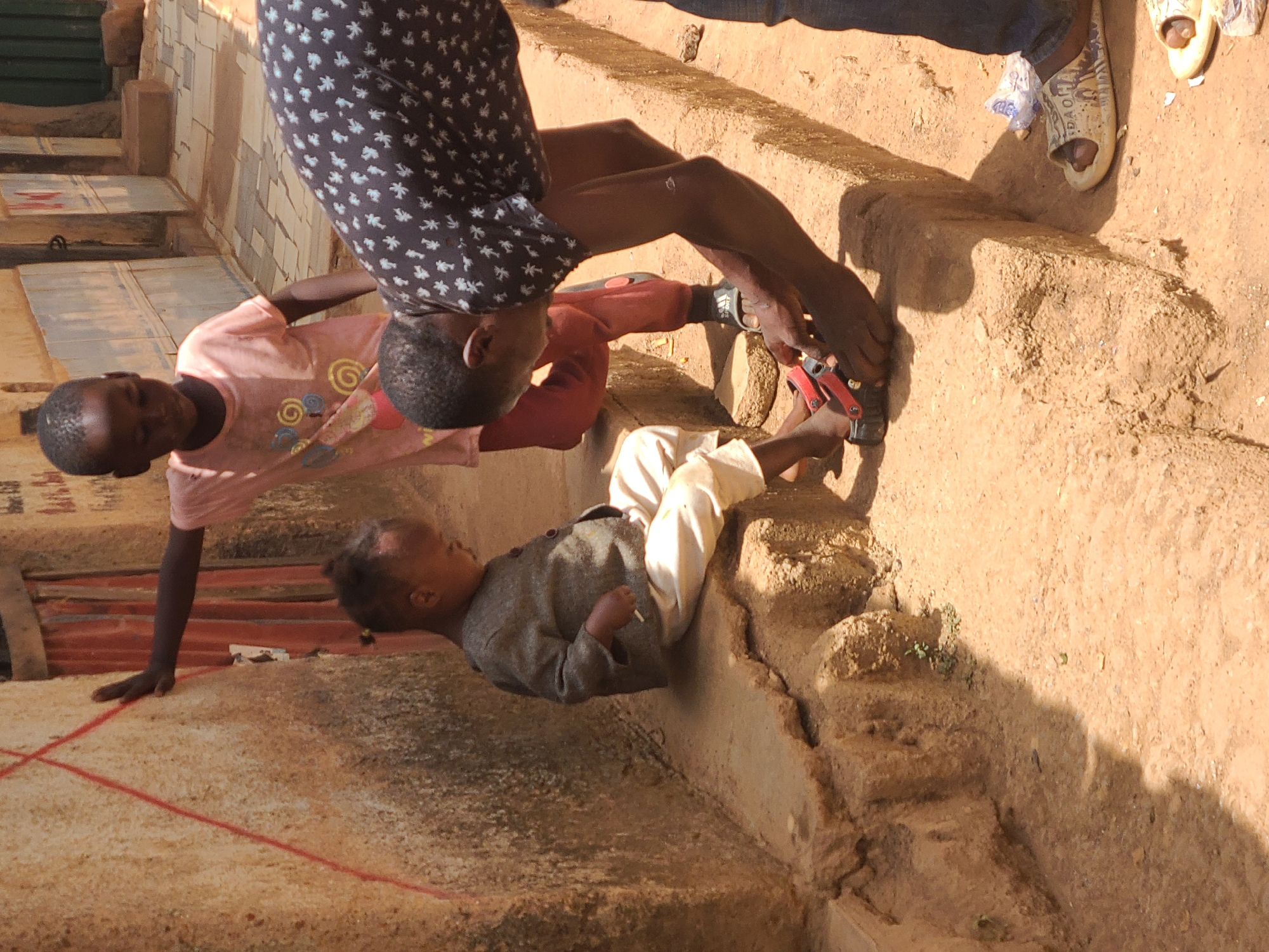 The image captures a scene in what appears to be a residential area of Jos, Nigeria. Three individuals are visible. A child wearing a pink shirt is in a handstand position, balancing with one hand on the ground and the other hand on the side of a concrete pathway. An older individual wearing a dark, patterned shirt is kneeling beside the child, appearing to assist or adjust the child's footwear. A third child in a gray shirt and white pants is lying down in the pathway to the side of the other two. The pathway is composed of soil and concrete, and it appears to be situated adjacent to a building with corrugated metal paneling and painted walls. Sandals and other debris are scattered on the ground nearby. The image is brightly lit, indicating daytime.