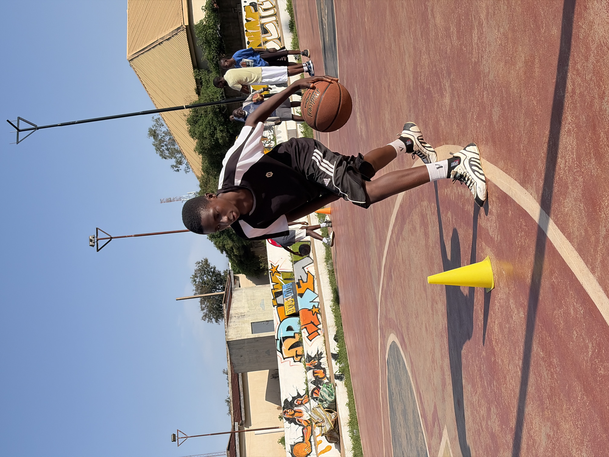 In Jos, Nigeria, a young Black male basketball player is practicing dribbling on an outdoor court. He is wearing a black and white t-shirt, black shorts with white stripes, and white and black sneakers with white socks. He is actively dribbling a basketball. In the background, other individuals are present, with a wall decorated with graffiti behind them. The court is made of a reddish-brown material with white lines. A yellow traffic cone is visible on the court. The sky is clear and blue, and there are buildings and trees in the distance.