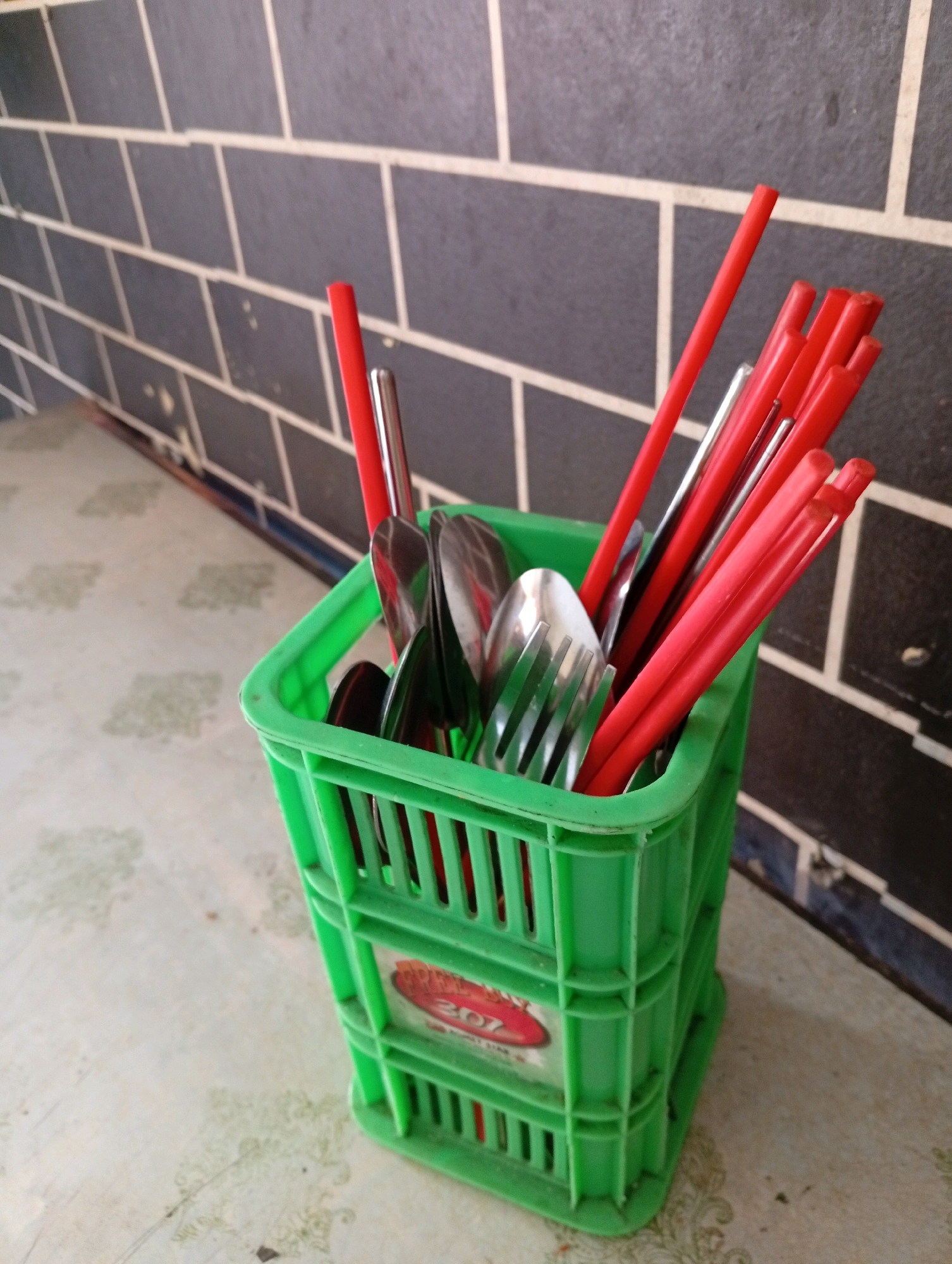 A close-up, eye-level shot shows a bright green plastic crate filled with eating utensils, including red chopsticks, metal spoons, and metal forks. The crate is positioned on a patterned surface. In the background, a dark gray brick wall with white grout lines is visible. The lighting appears to be natural, casting soft shadows. This scene is likely from East Jakarta, Indonesia, given the common use of such crates and utensils in local eateries or street food stalls.