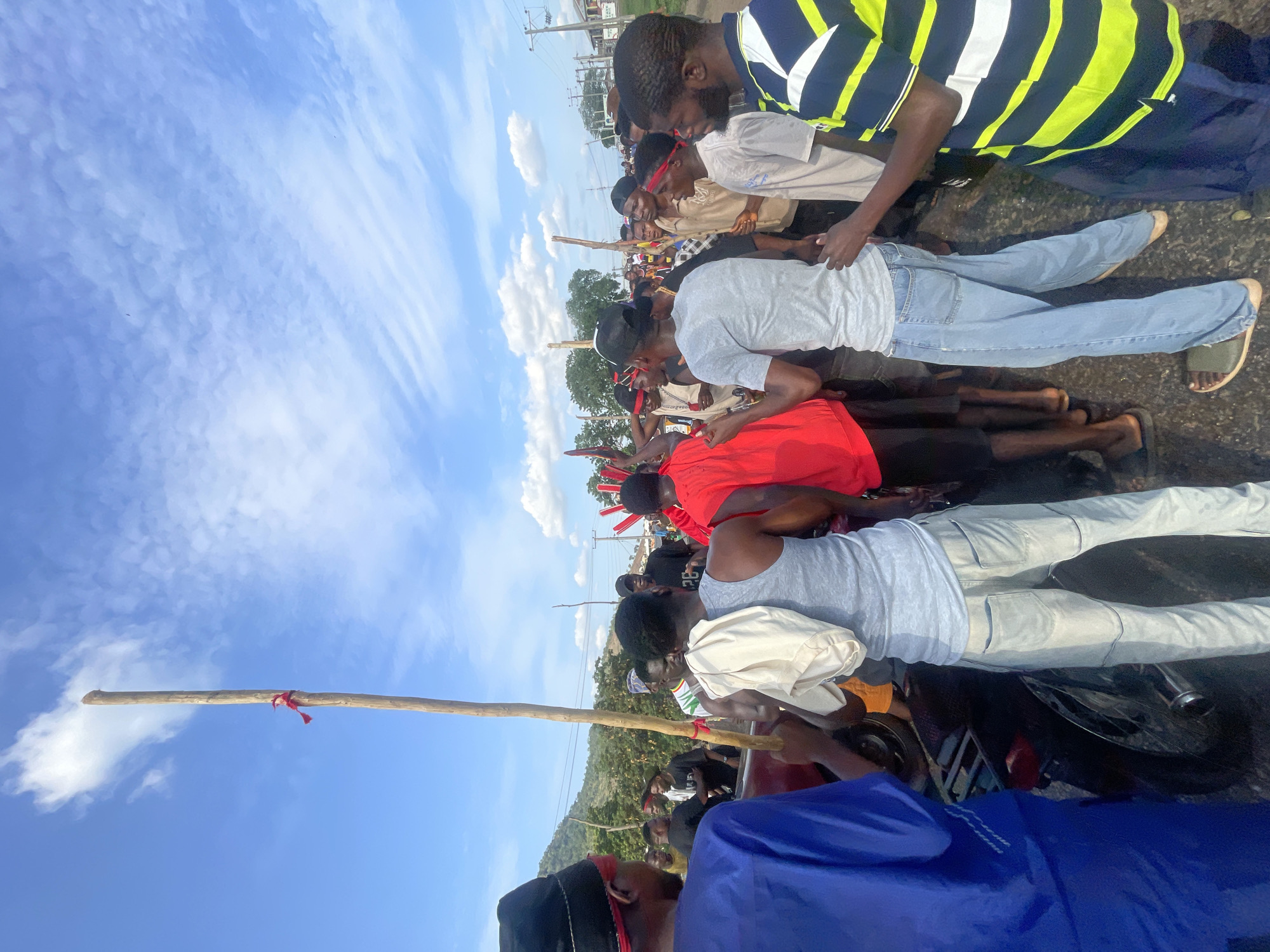 A horizontal, medium shot captures a group of people, primarily young men, standing along a road in Lokoja, Nigeria. The sky is visible in the background, featuring a blue hue with scattered white clouds. Some individuals are wearing red headbands and a few are holding wooden poles, with one pole having a red ribbon tied to it. Their clothing varies, including t-shirts (gray, red), a striped shirt, and a blue garment. The individuals are standing on asphalt and a motorcycle is present. There are utility poles and sparse vegetation in the background.