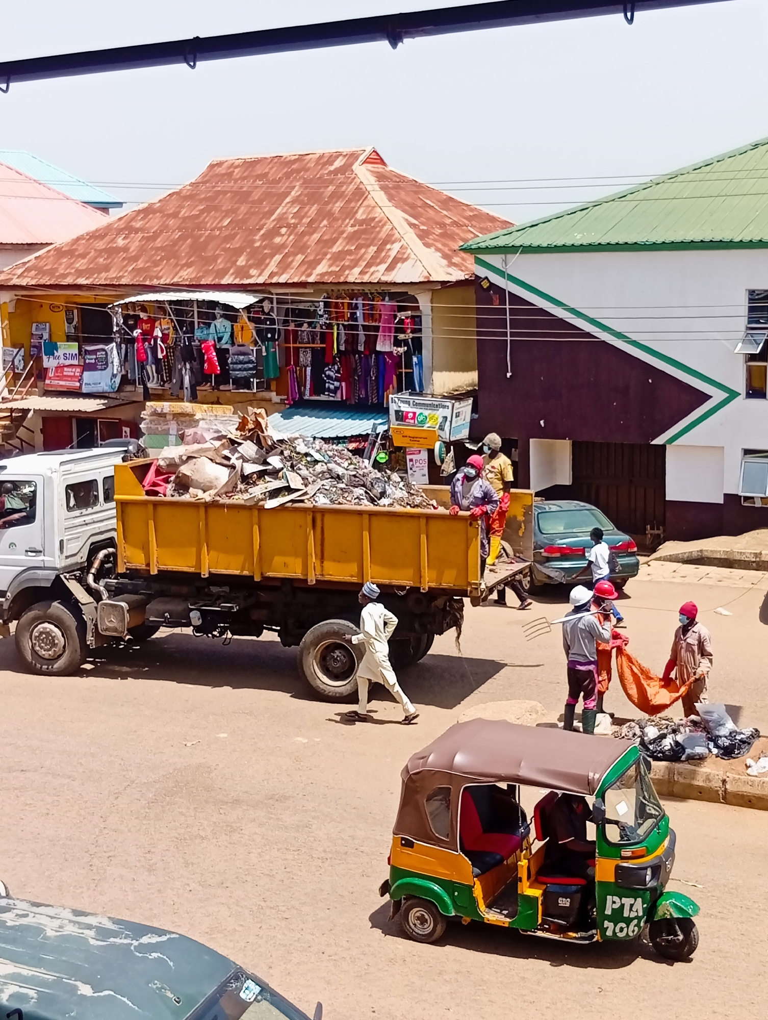 In Bukuru, Nigeria, on a clear day, a scene unfolds by a row of shops with corrugated iron roofs, one rusty brown and the other green. A large, yellow dump truck, laden with debris and what appears to be construction waste, is parked on a dusty road. Several individuals are engaged in the activity of clearing or loading the truck.
On the left, the driver's side of the truck is visible, with a figure in the driver's seat. A man in a light-colored outfit and a cap walks away from the truck towards the right.
In the center, near the back of the truck, a group of people are actively involved in waste management. Some are wearing masks and gloves, suggesting safety precautions. One individual, dressed in work clothes and a red hard hat, holds a pitchfork. Another person in orange overalls and a red head covering is seen holding a large orange tarp or bag. Next to them, a pile of clothing or fabric is laid out on the ground. A dark-colored sedan is parked behind the truck.
A young boy in a white shirt and dark shorts is walking near the sedan. In the foreground, a colorful auto-rickshaw, green, yellow, and brown, with the license plate PTA 706, is parked facing left. Another vehicle, only partially visible, is parked in the lower left corner of the image. The buildings behind the truck appear to be shops, with clothing displayed on racks outside some of them.