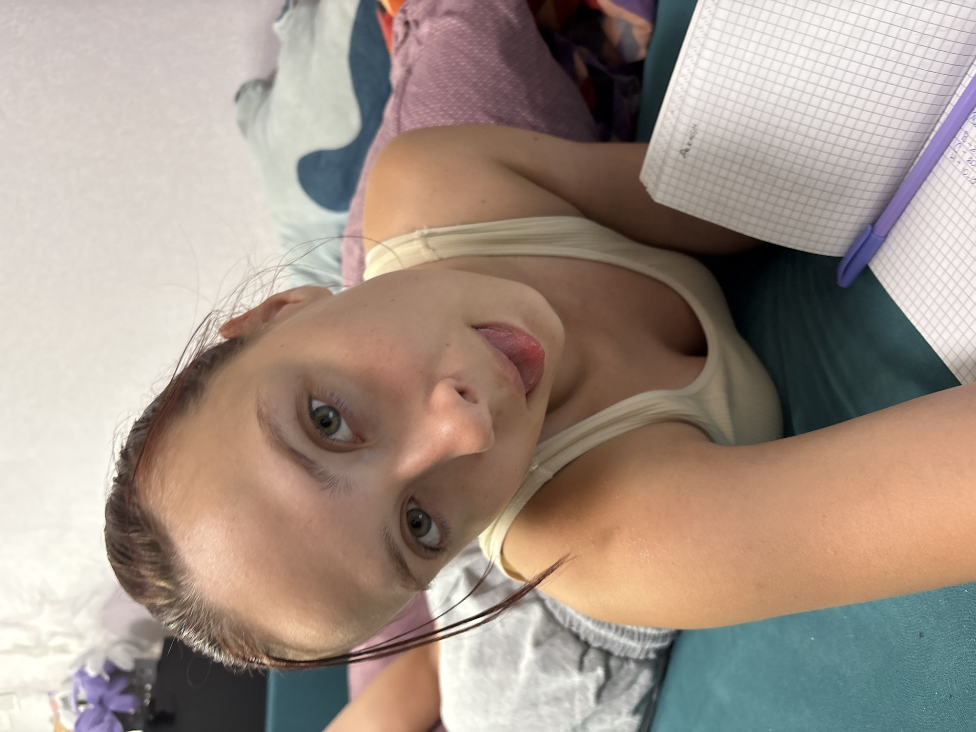 A young female, appearing to be in her late teens or early twenties, is depicted in a close-up, low-angle shot, lying prone on a teal-colored surface in the city of Прикамский, Russia. Her face is centered, featuring green eyes and a fair complexion. Her hair, appearing dark brown with reddish highlights and slightly wet or slicked back, frames her face. Her mouth is slightly open, with her tongue partially extended, conveying a playful expression. She is wearing a light-colored, possibly cream or pale yellow, ribbed tank top, and grey sweat shorts are visible on her lower body. In the foreground to the bottom right, an open grid-lined notebook with some handwritten notes and a purple pen resting upon it are visible. The background reveals various soft furnishings, including purple and light blue textiles with a dark blue abstract shape, suggesting a bed or couch. In the top left background, artificial purple flowers in a dark container are partially visible against a light-colored wall. The scene is illuminated by soft, even lighting, indicating an indoor setting.