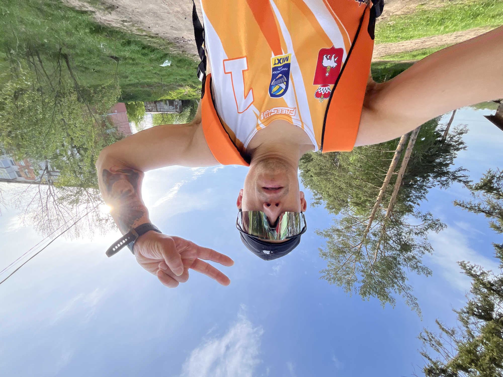 A man wearing sunglasses, a black cap, and an orange and white athletic vest with the number "1" and logos, including one possibly of Russian origin, is taking a selfie while making a peace sign. The background is a bright blue sky with scattered clouds and the tops of trees. Some grass and a building are visible in the top right. This is most likely outdoors and during the day. The city of Chaikovsky, Russia is not conclusively discernible.
