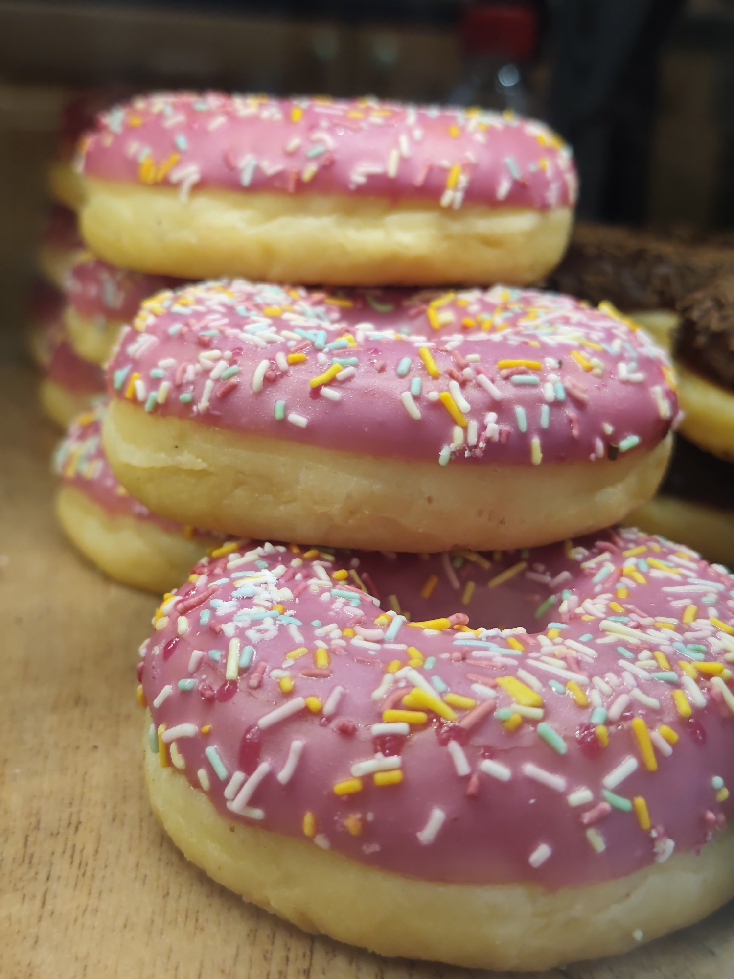 A close-up, eye-level shot shows a stack of donuts on a wooden surface. The donuts are round with a hole in the center, covered in pink icing and sprinkled with colorful, thin, rod-shaped sprinkles in white, pink, yellow, and light blue. The donuts are stacked in a staggered, slightly diagonal fashion, with the closest donut filling the bottom right of the frame. The lighting is soft, highlighting the texture of the icing and sprinkles. The background is blurred and dark, with hints of other objects, possibly more food items, and some red and white lights. The image does not provide any information or context to determine its location in Sochi, Russia.
