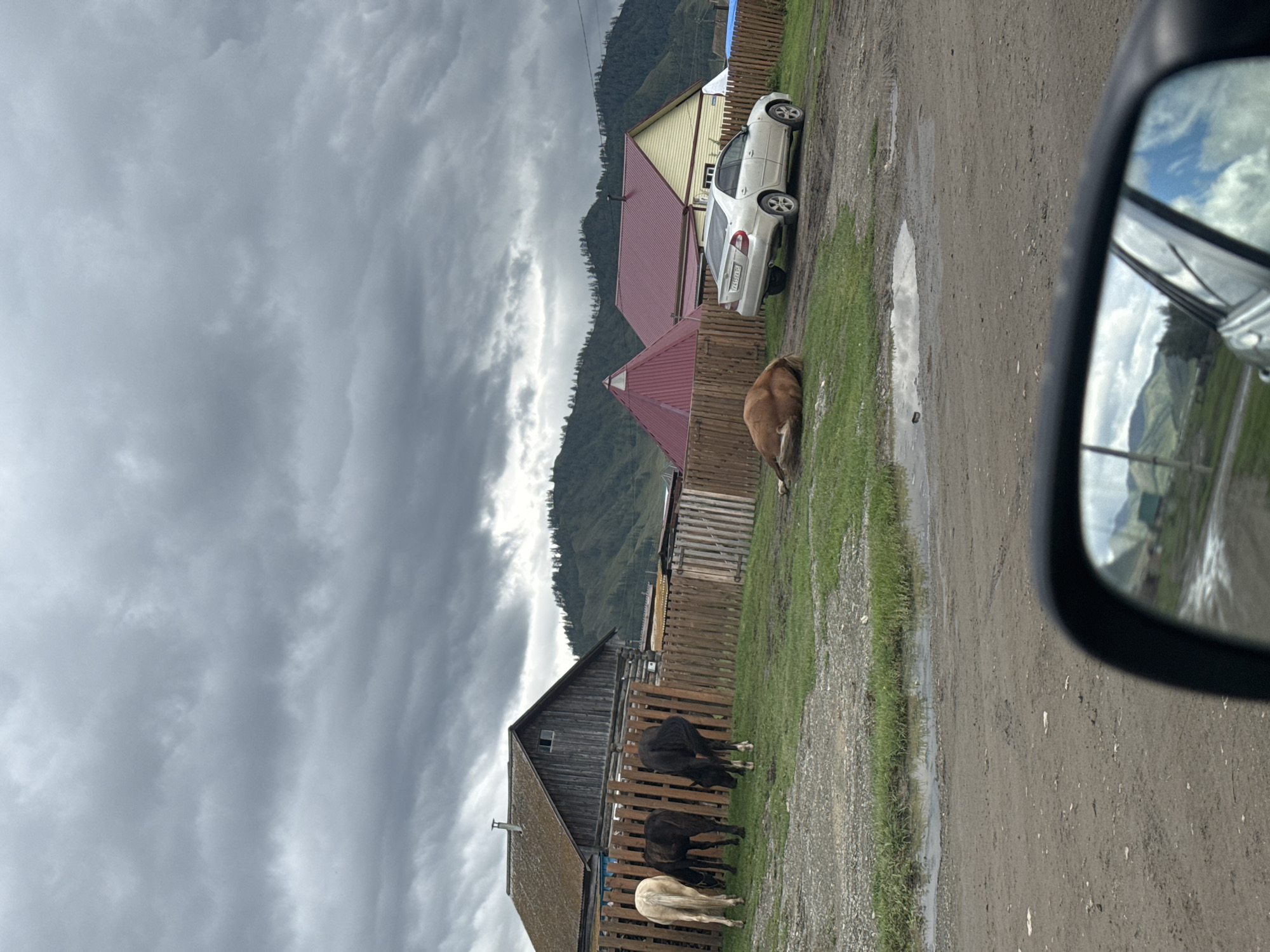 The image depicts a rural scene in Yakonur, Russia, on a cloudy day. In the foreground, a dirt road with puddles is visible, and a car's side mirror reflects a cloudy sky and rolling hills. A light-colored sedan is parked on the side of the road. To the left, three horses are grazing in a grassy enclosure behind a wooden fence. One horse, lying down on the grass, appears to be sleeping or resting. Behind the fence, several wooden houses and structures with pitched roofs are situated. The background features a range of tree-covered hills under a dramatic, overcast sky.