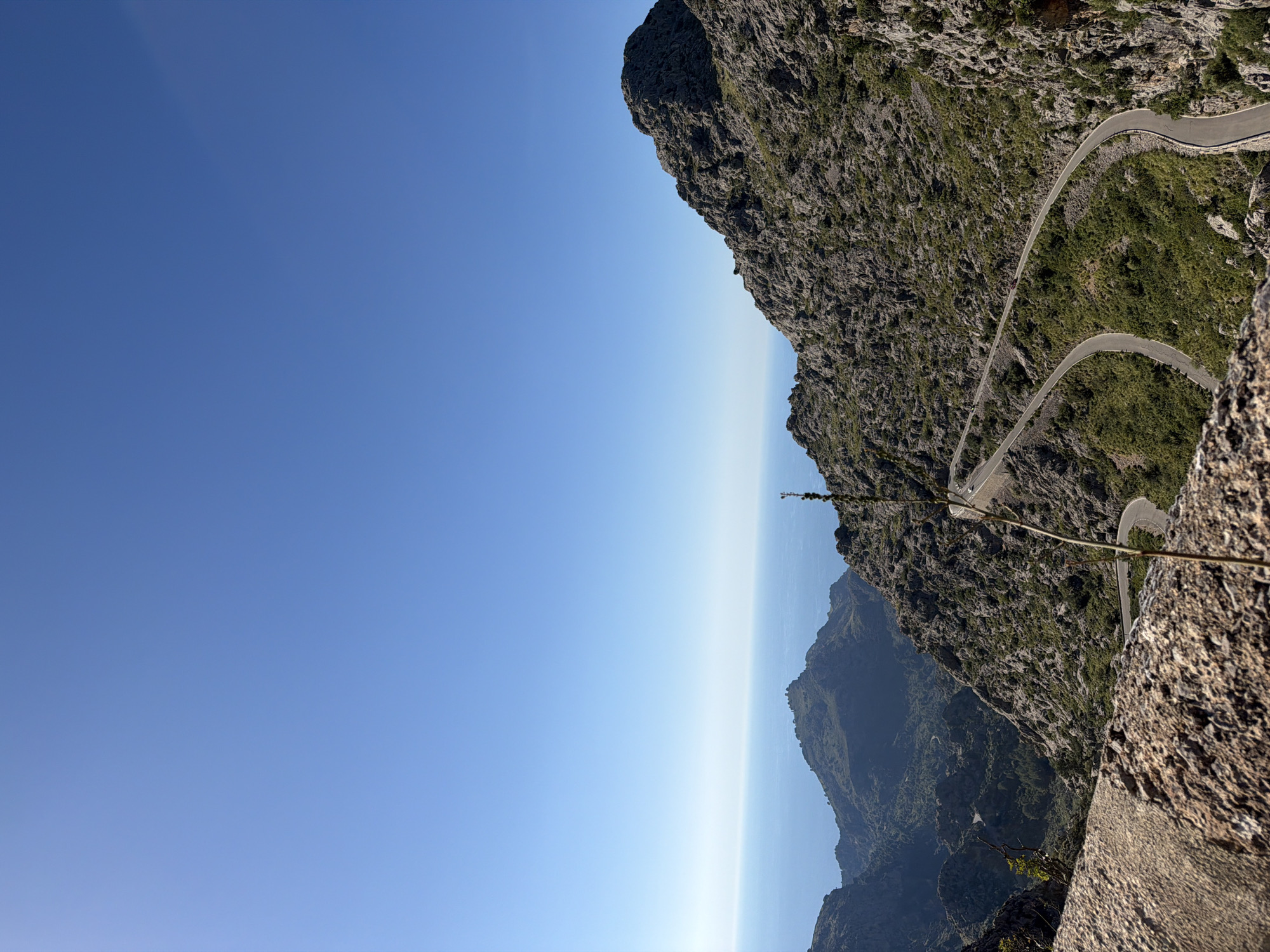 The image depicts a mountainous landscape in Escorca, Spain, under a clear blue sky. A winding road clings to the side of a rugged mountain. The road has sharp hairpin turns. The foreground features a rocky outcrop, and the background reveals a hazy view of the Mediterranean Sea. The overall scene conveys a sense of elevation and scenic beauty.