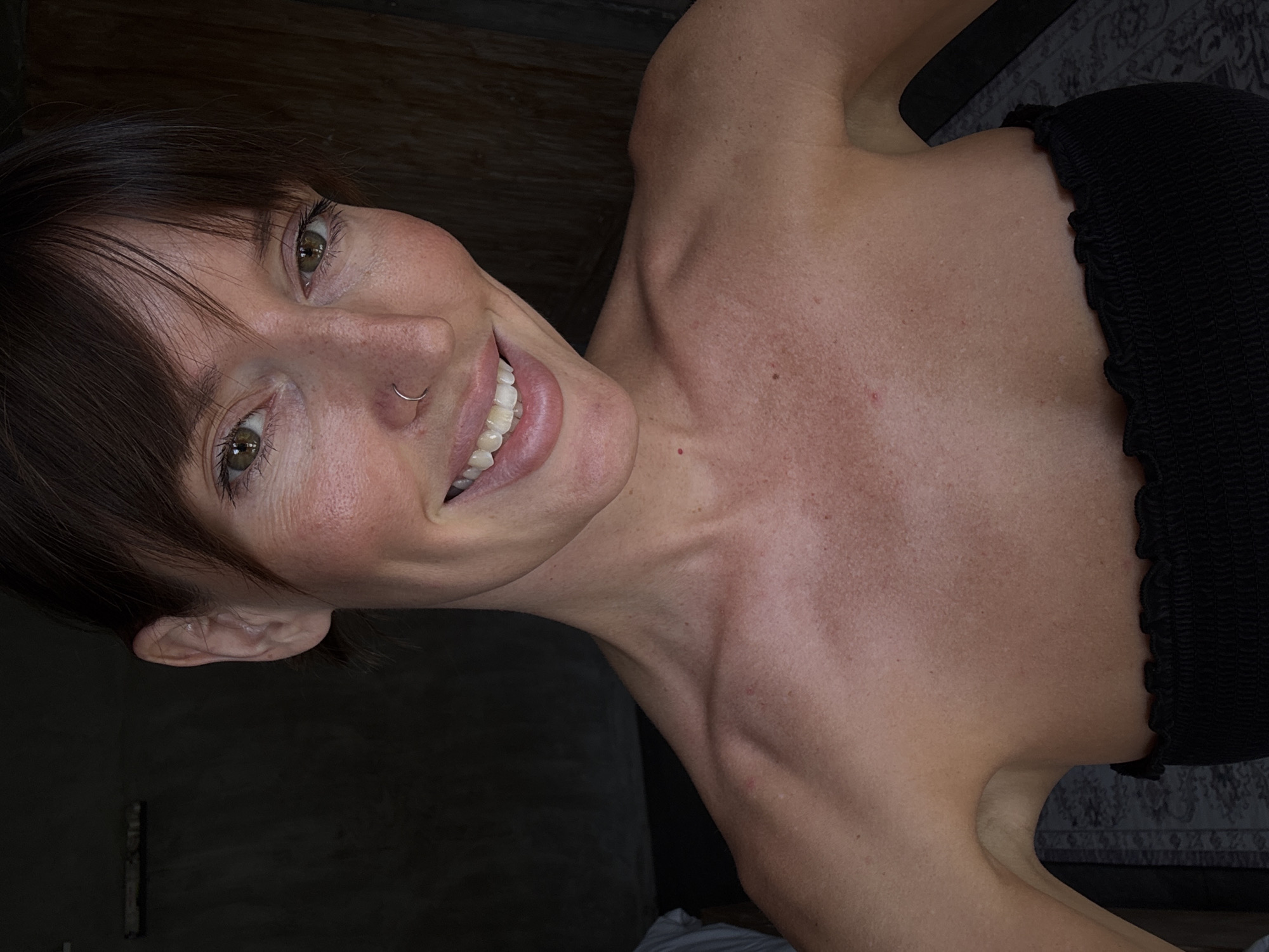 A woman with brown hair and a nose ring is smiling at the camera. She is wearing a black strapless top. The background is dark, with a wall visible on the left and a patterned rug on the right. The location is not discernible in the image to be North Kuta, Indonesia.