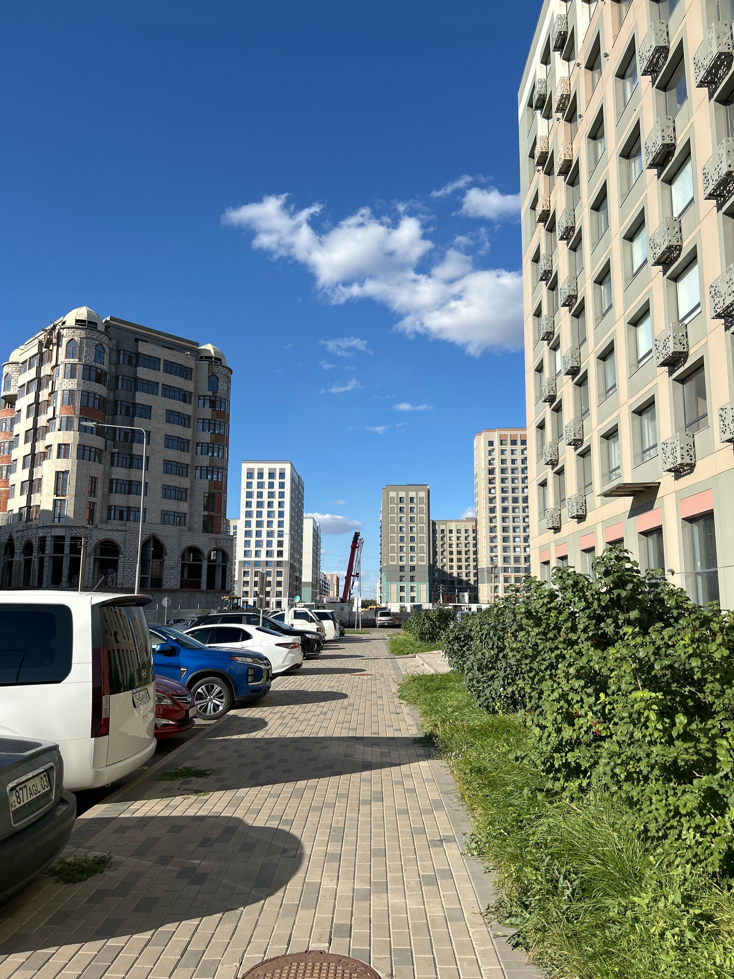 The image depicts a street scene in Astana, Kazakhstan, featuring modern residential buildings and parked vehicles. The architecture is contemporary, with multi-story buildings characterized by modular designs and prominent balconies. A row of parked cars, including a white van, a blue sedan, and other vehicles, lines a paved walkway. Lush green bushes and grass flank the walkway, contributing to the urban landscaping. The sky is a clear blue with scattered white clouds, indicating fair weather. A construction crane is visible in the background, suggesting ongoing development in the area. The overall impression is one of a developing urban environment with a focus on modern residential construction.