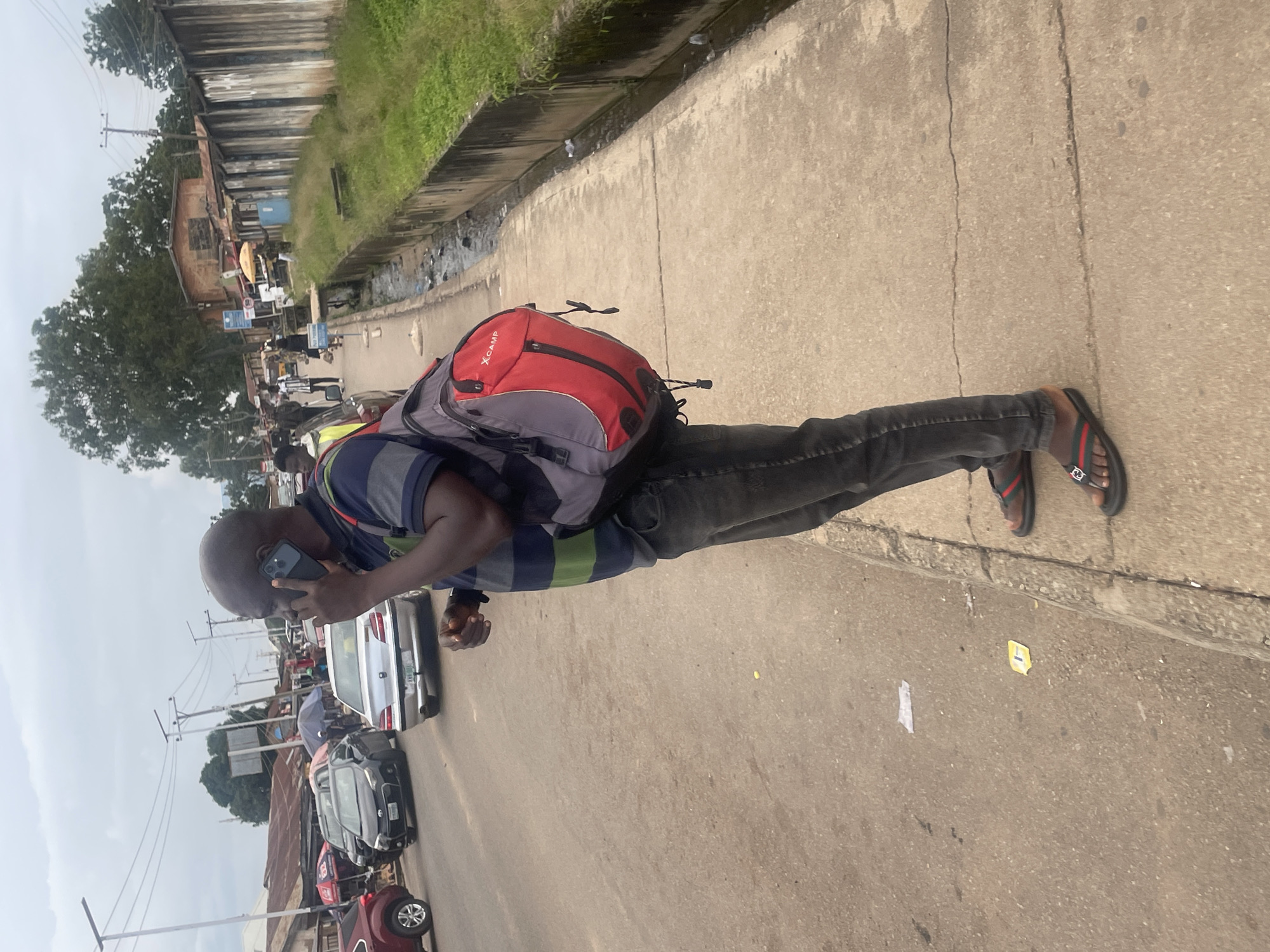 A man stands on a concrete sidewalk next to a road in Jos, Nigeria, speaking on his phone. He wears a striped t-shirt, dark jeans, and flip-flops. A red and gray backpack is slung over his right shoulder. The road is busy with traffic, including cars and what appears to be a motorcycle. Buildings and market stalls line the opposite side of the road. A concrete drainage ditch runs alongside the sidewalk. The sky is overcast.